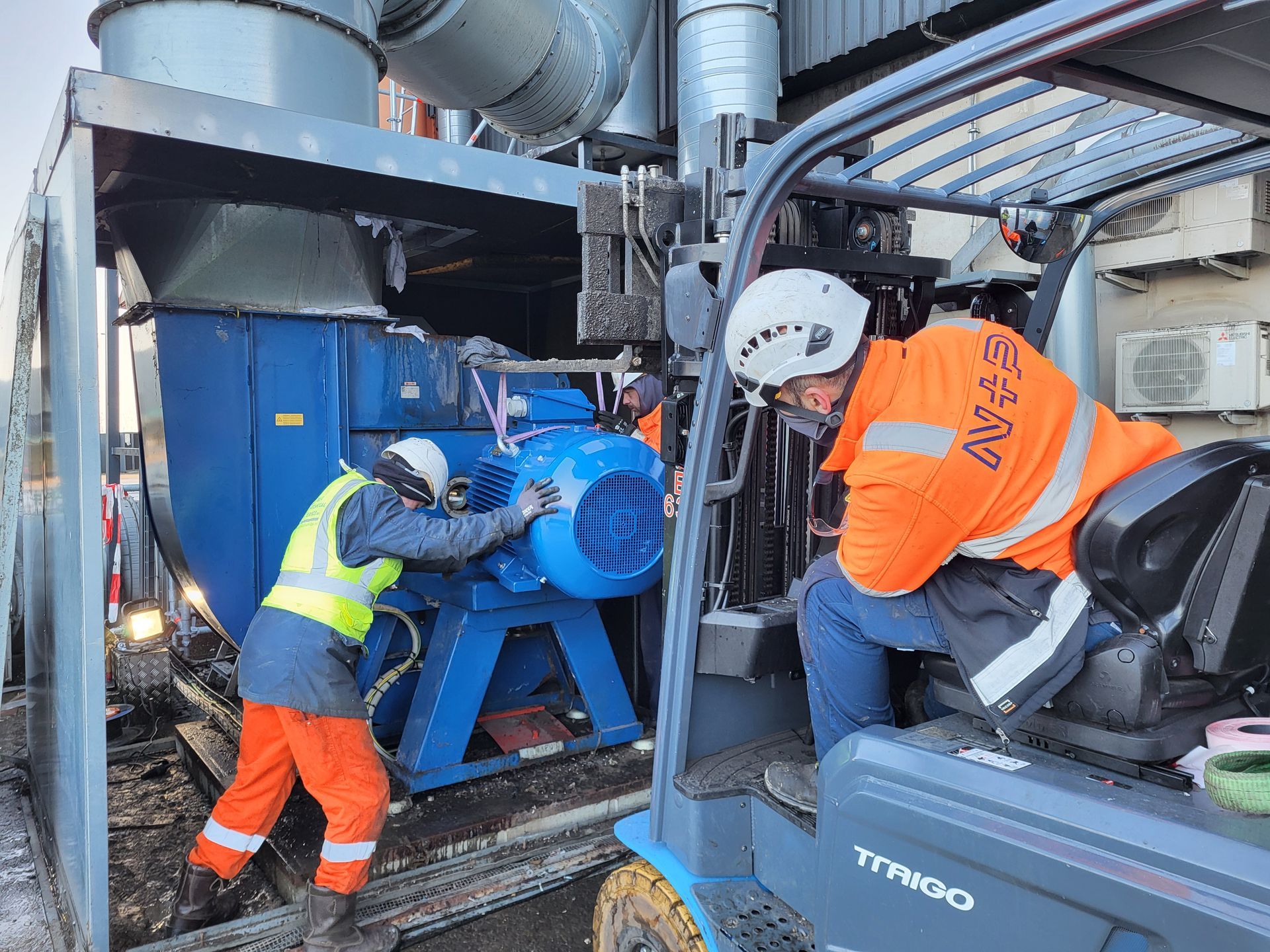 Twee arbeiders installeren buiten een groot blauw machineonderdeel met behulp van een heftruck.