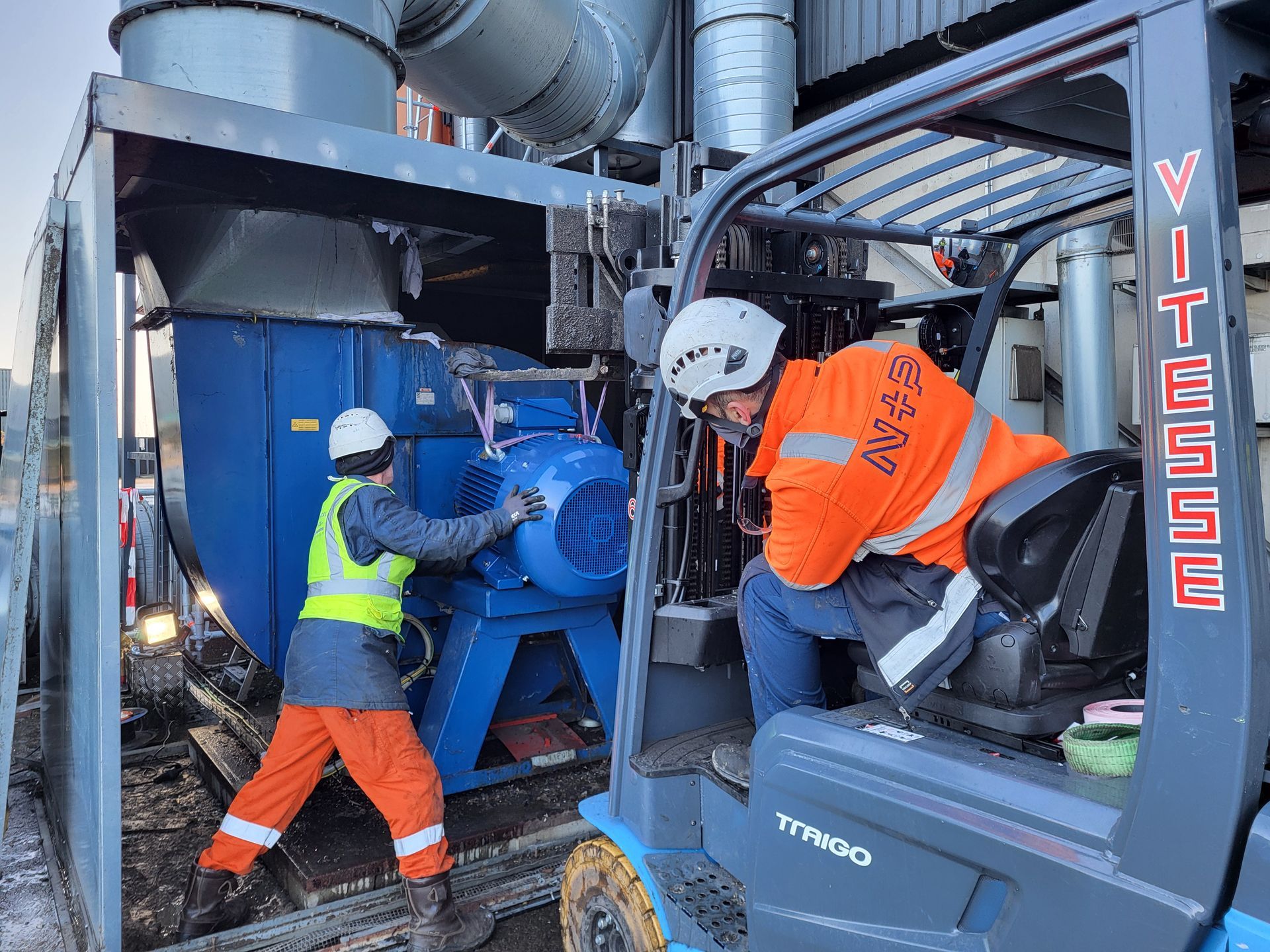 Twee arbeiders installeren blauwe industriële apparatuur, waarvan één op een heftruck, in de buitenlucht.