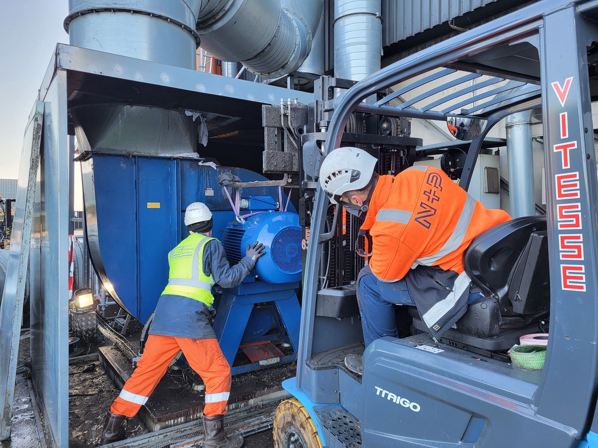 Arbeiders installeren blauwe industriële apparatuur, één van hen zit in een heftruck.