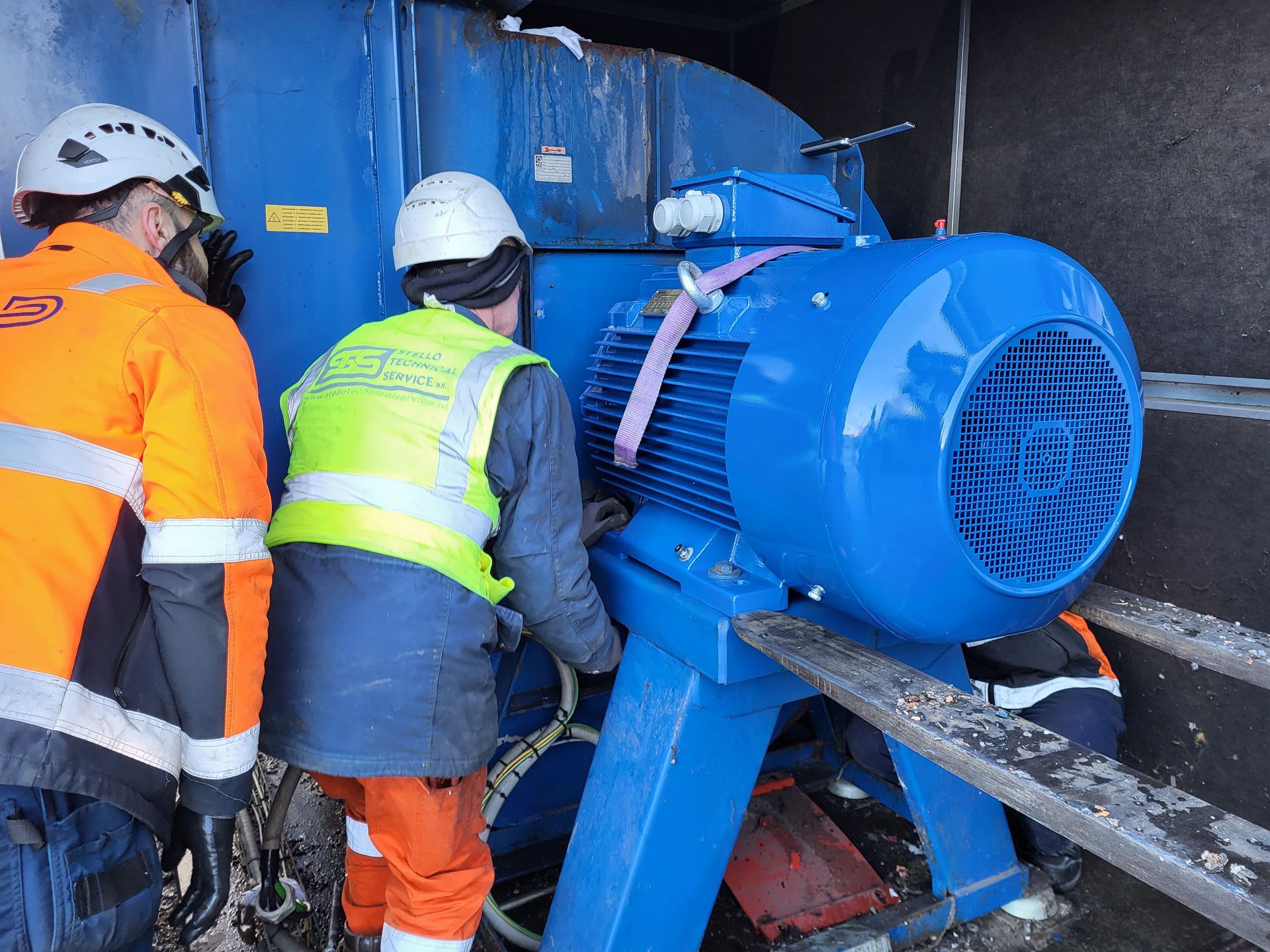 Arbeiders in veiligheidsuitrusting verplaatsen een grote blauwe industriële motor met behulp van een takel.