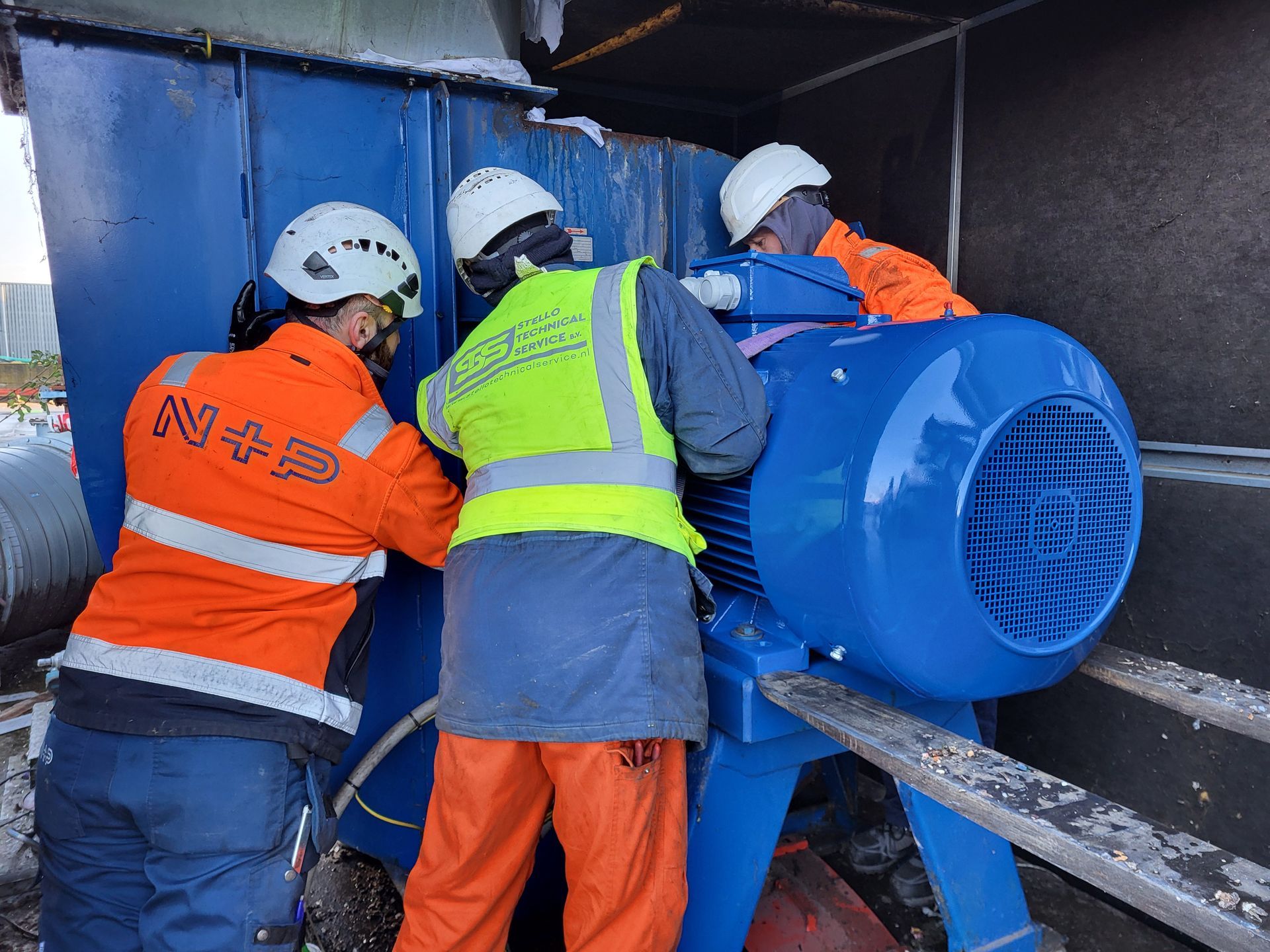 Drie werknemers in veiligheidsuitrusting installeren een grote blauwe motor.