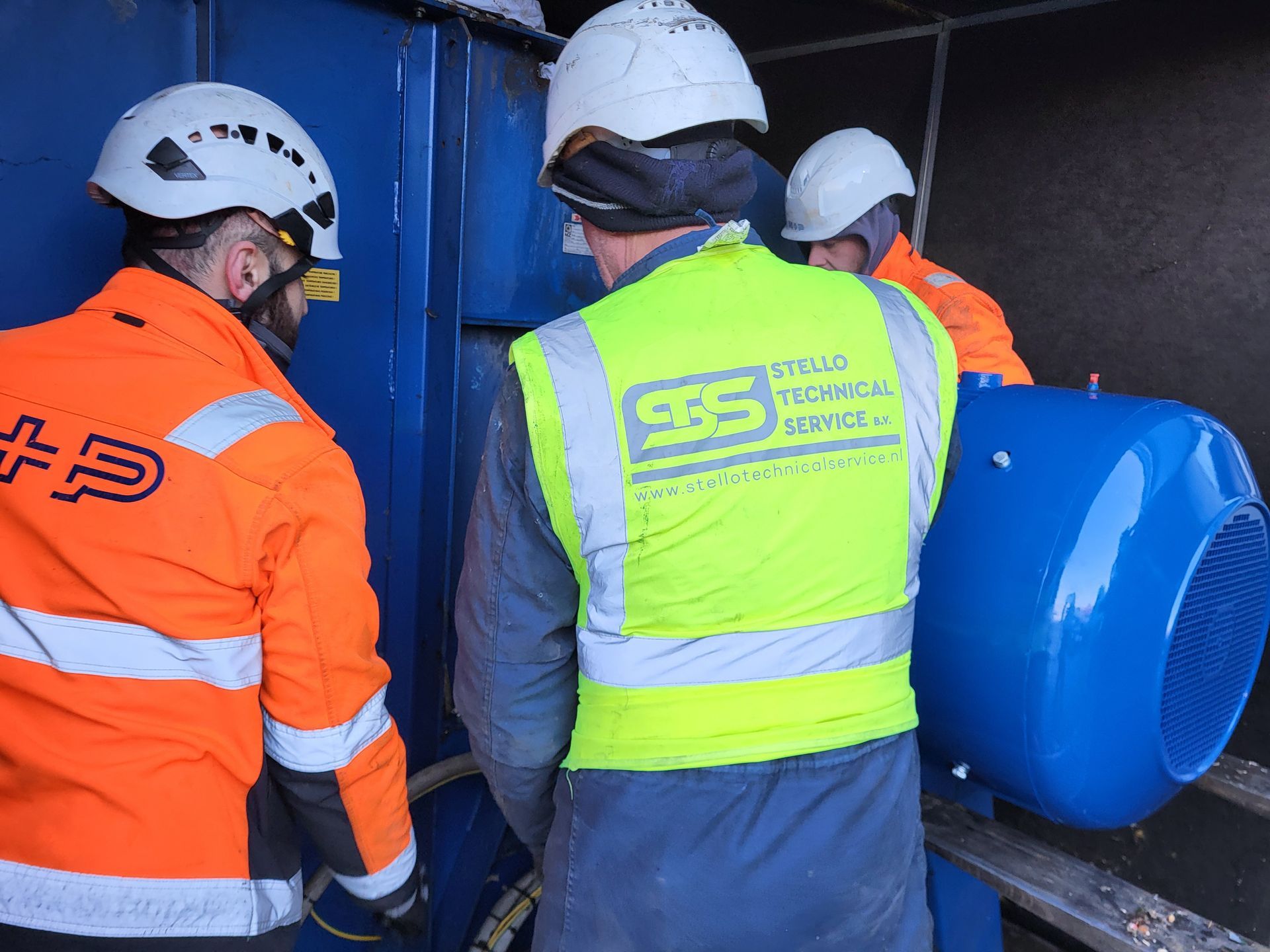 Drie werknemers in veiligheidsuitrusting installeren een blauw industrieel onderdeel.