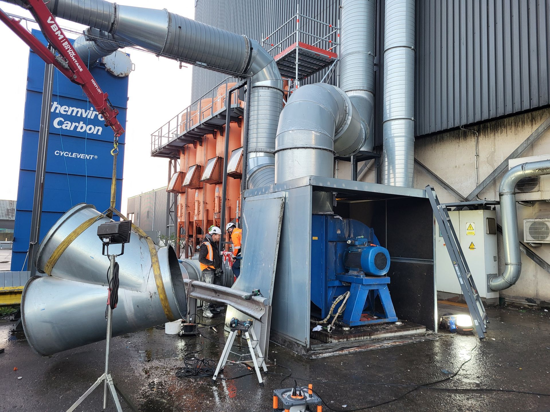 Industriële machines met werknemers in veiligheidsvesten, nabij een grote blauw-oranje constructie.