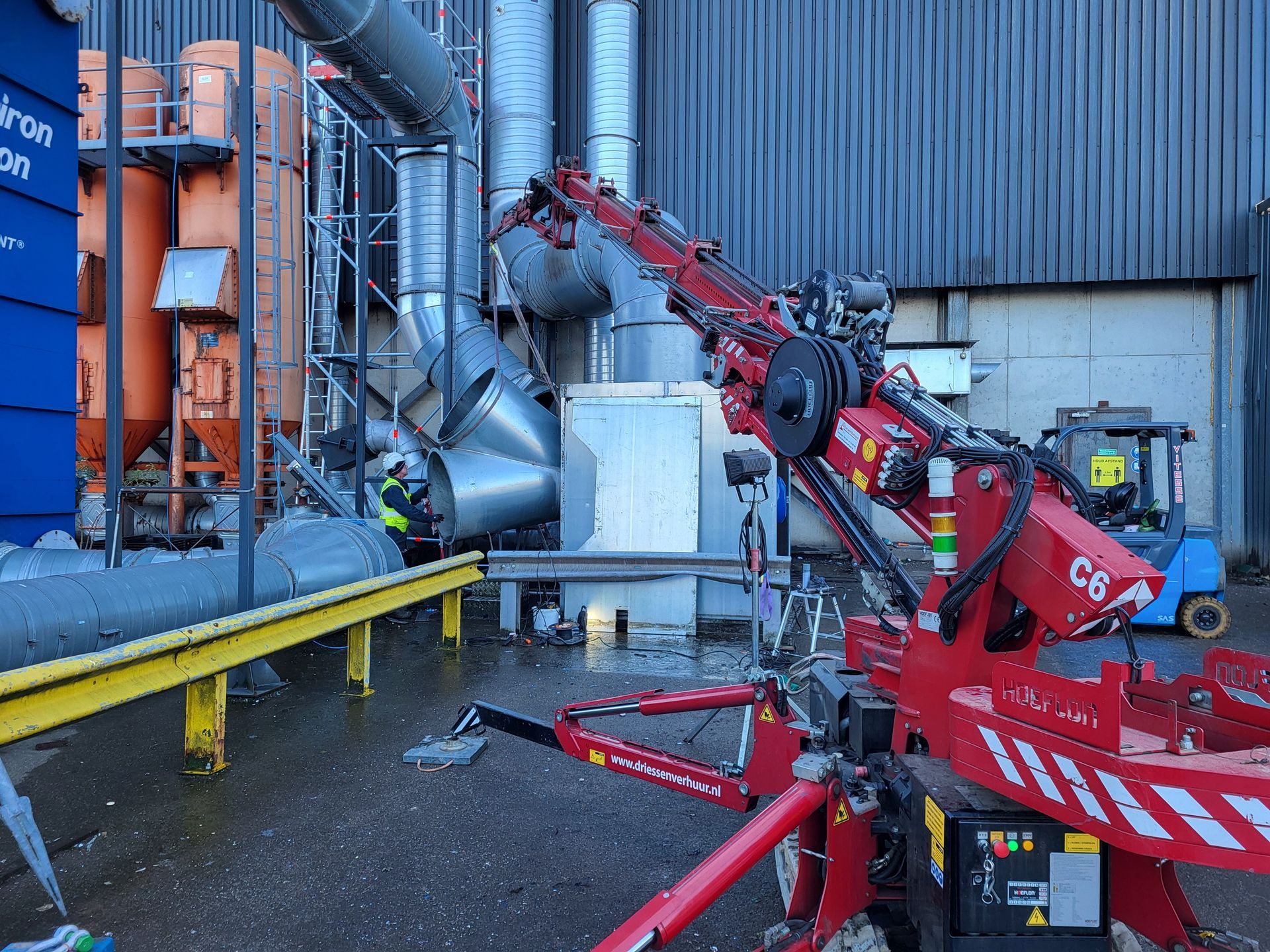 Rode kraan naast beschadigde industriële apparatuur buiten een blauw gebouw.