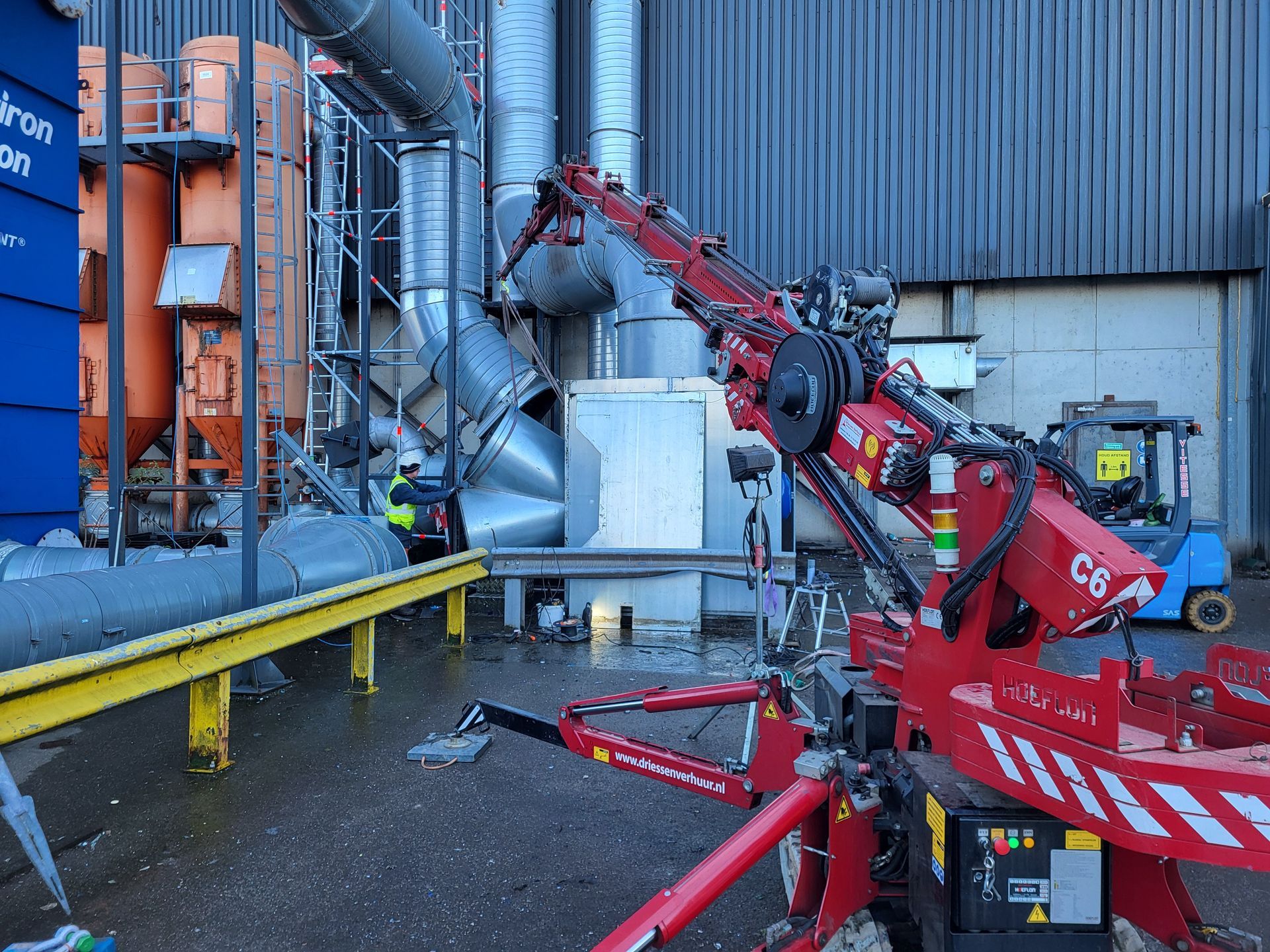 Een rode kraan demonteert industriële apparatuur in de openlucht, met een werknemer erbij.