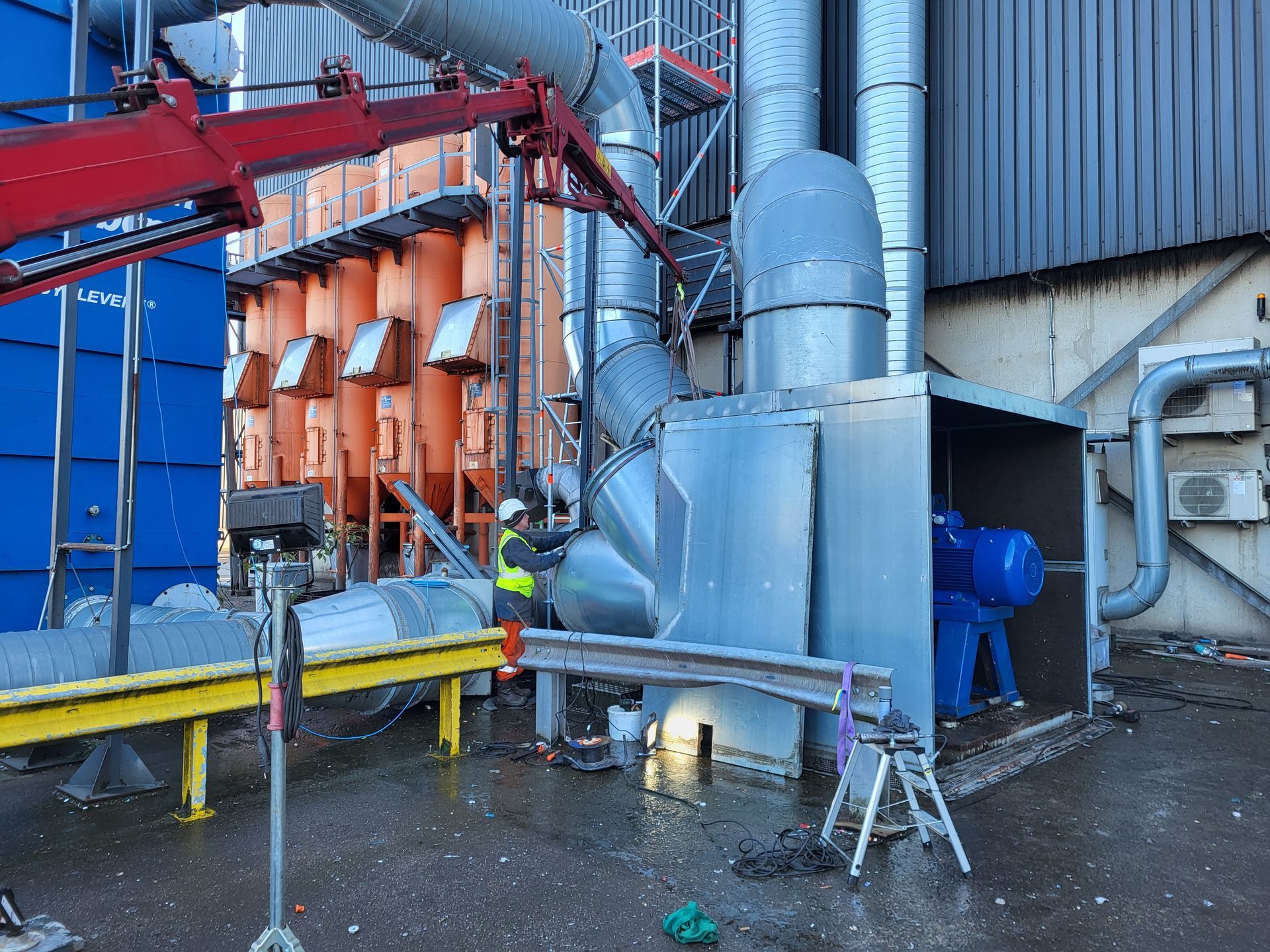 Arbeiders installeren industriële apparatuur; oranje en blauwe constructies, metalen buizen, kraan.