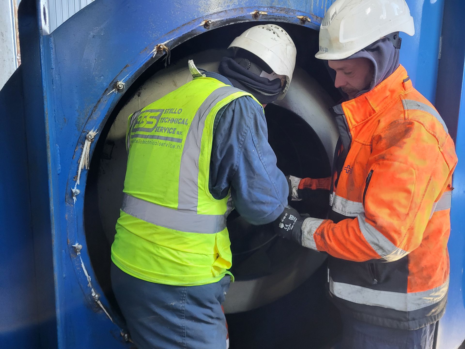 Twee werknemers in veiligheidskleding inspecteren machines. De ene draagt een geel vest, de andere een oranje jas.