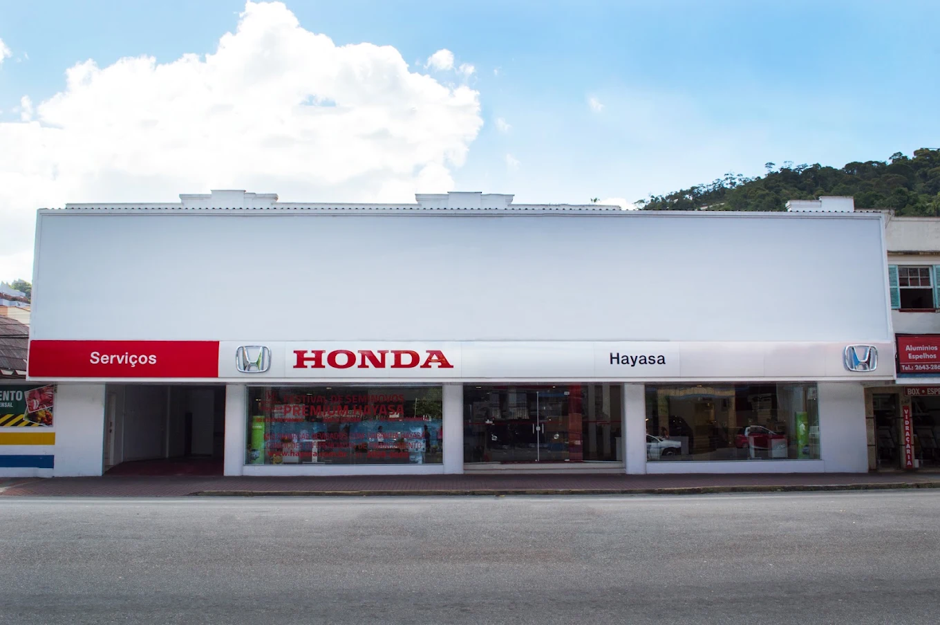 Fachada de um prédio, concessionária de automóveis. Carros estacionados do lado de fora. Céu azul.