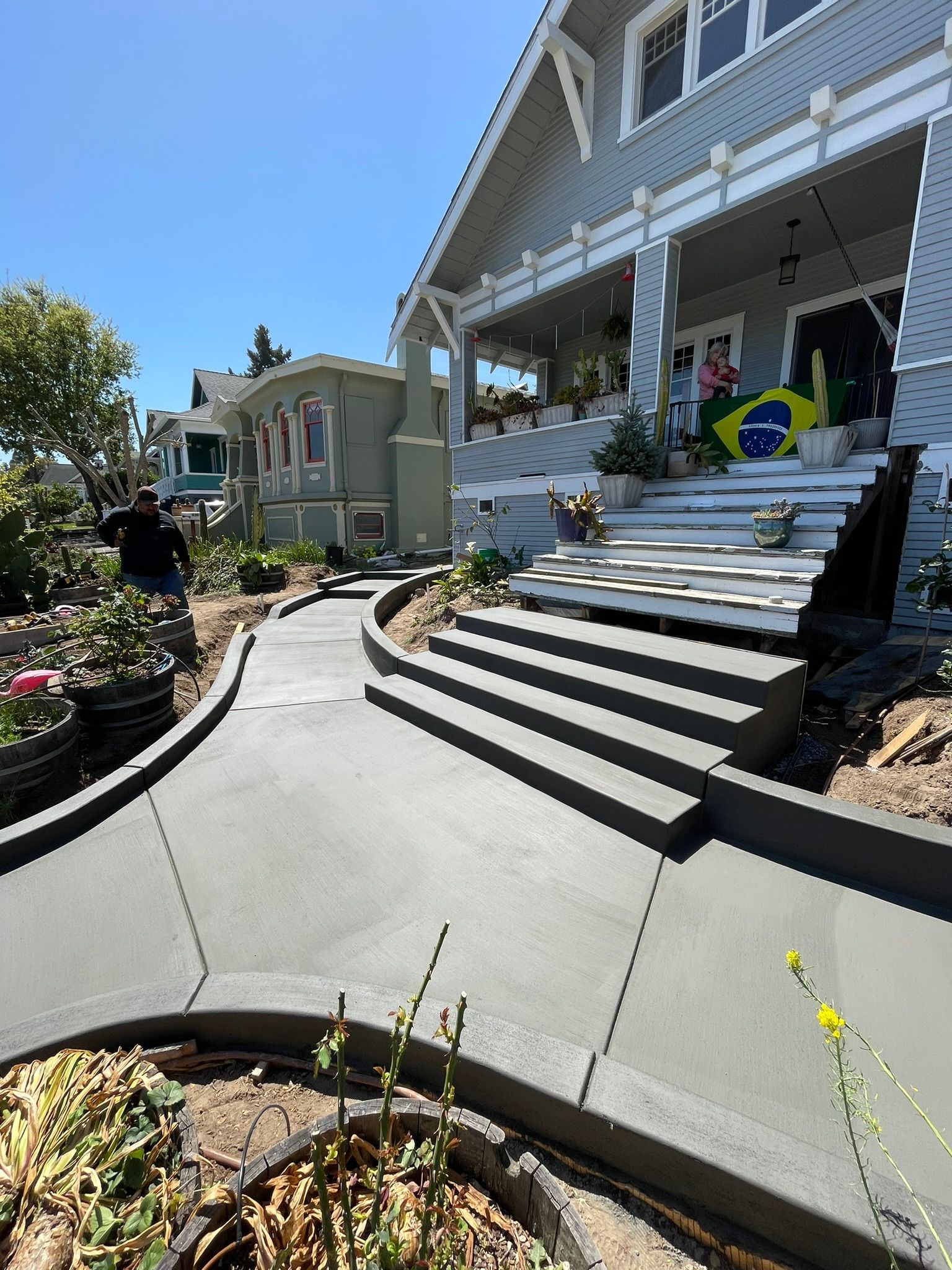 Steep white house steps with porch, flower beds, and a person near a Brazilian flag in bright sunlight