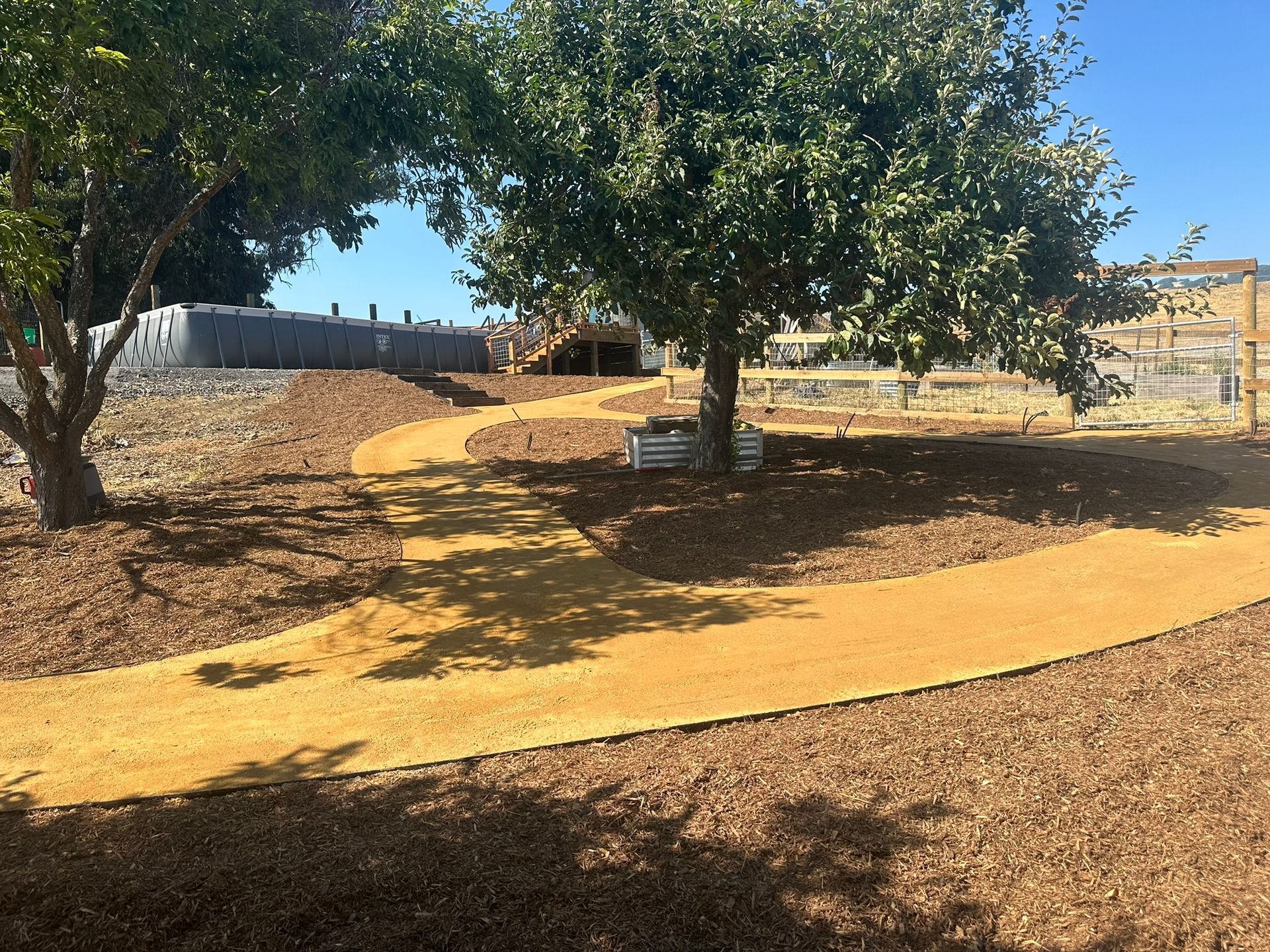 Curving yellow walking path through a park with trees and mulch under a clear blue sky
