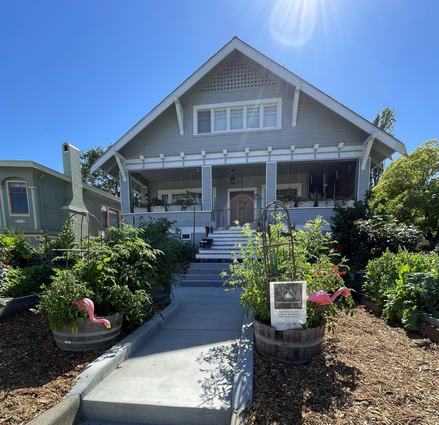 Front view of a gray house with a white porch, central steps, and landscaped garden under a sunny blue sky