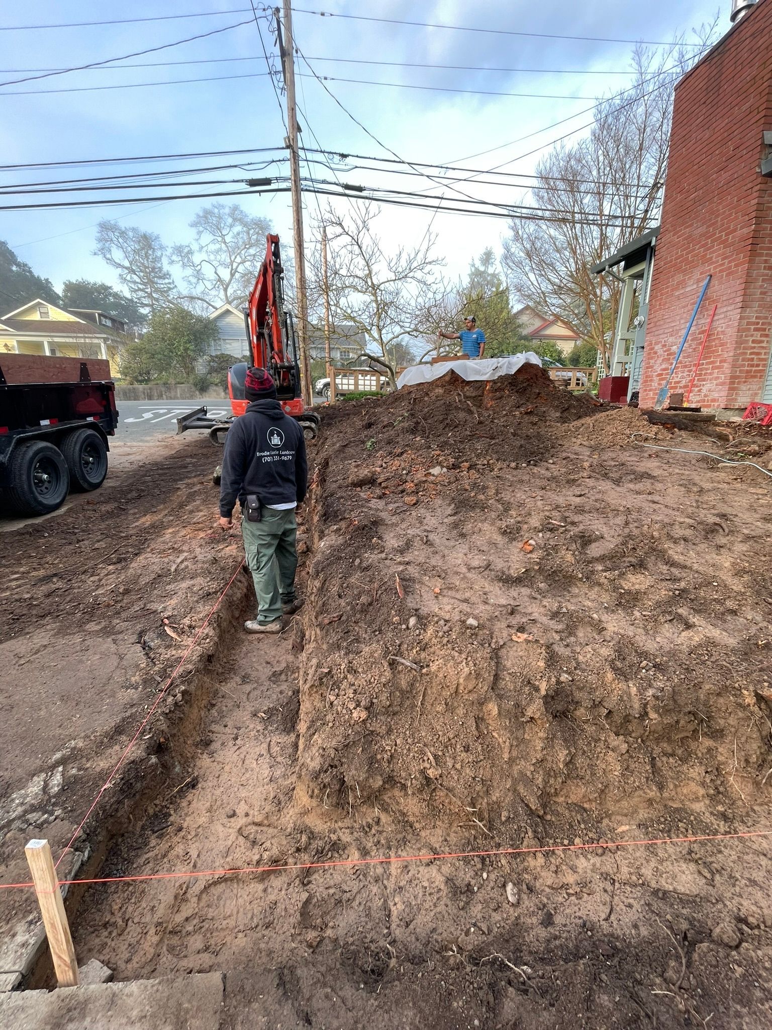 Worker beside excavator on muddy construction site next to a brick building