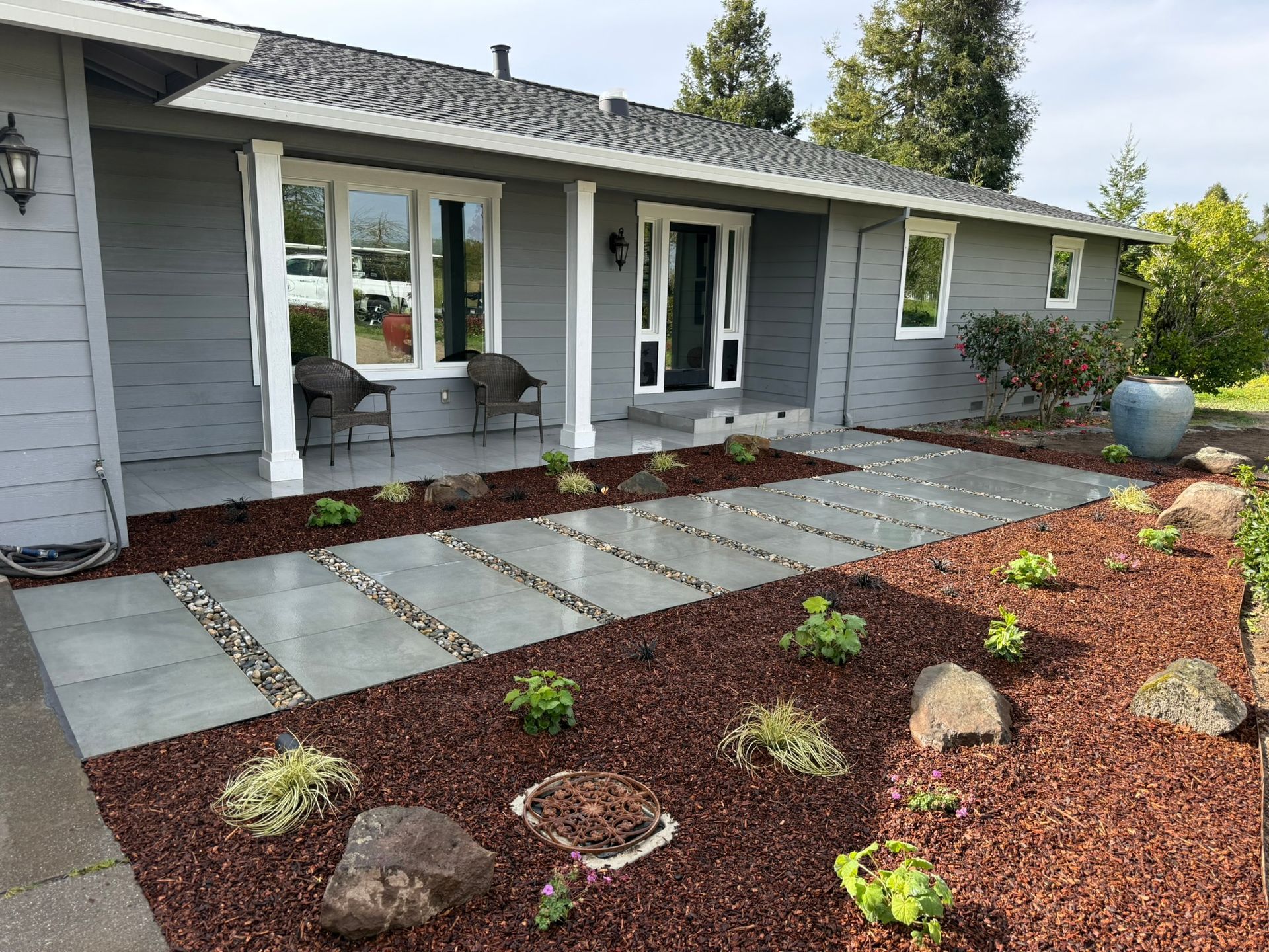 Front yard of a gray house with a stone patio, two chairs, and a landscaped path with rocks and mulch.