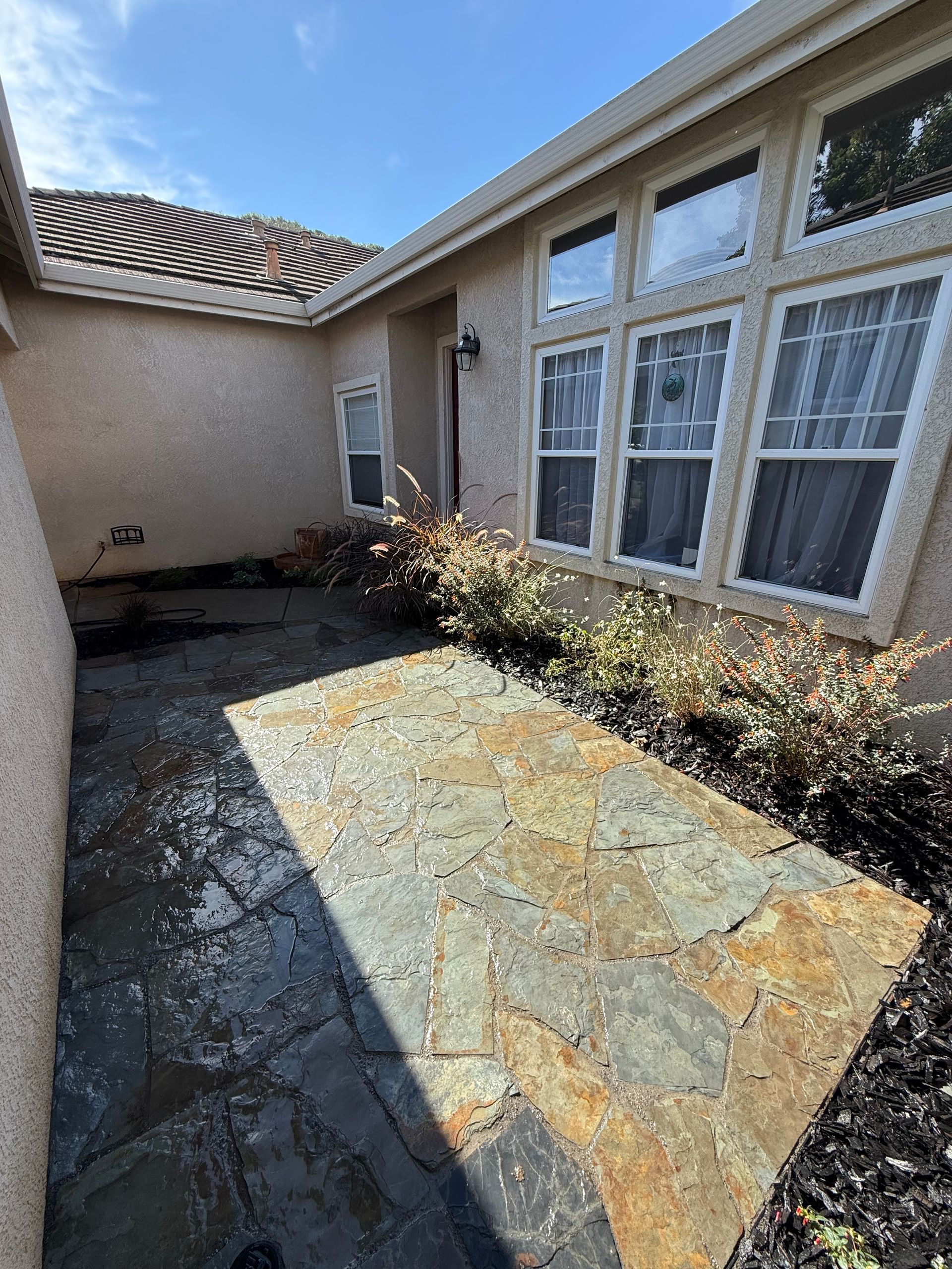 Sunny patio with stone pavers, beige stucco walls, and large windows beside a narrow garden bed