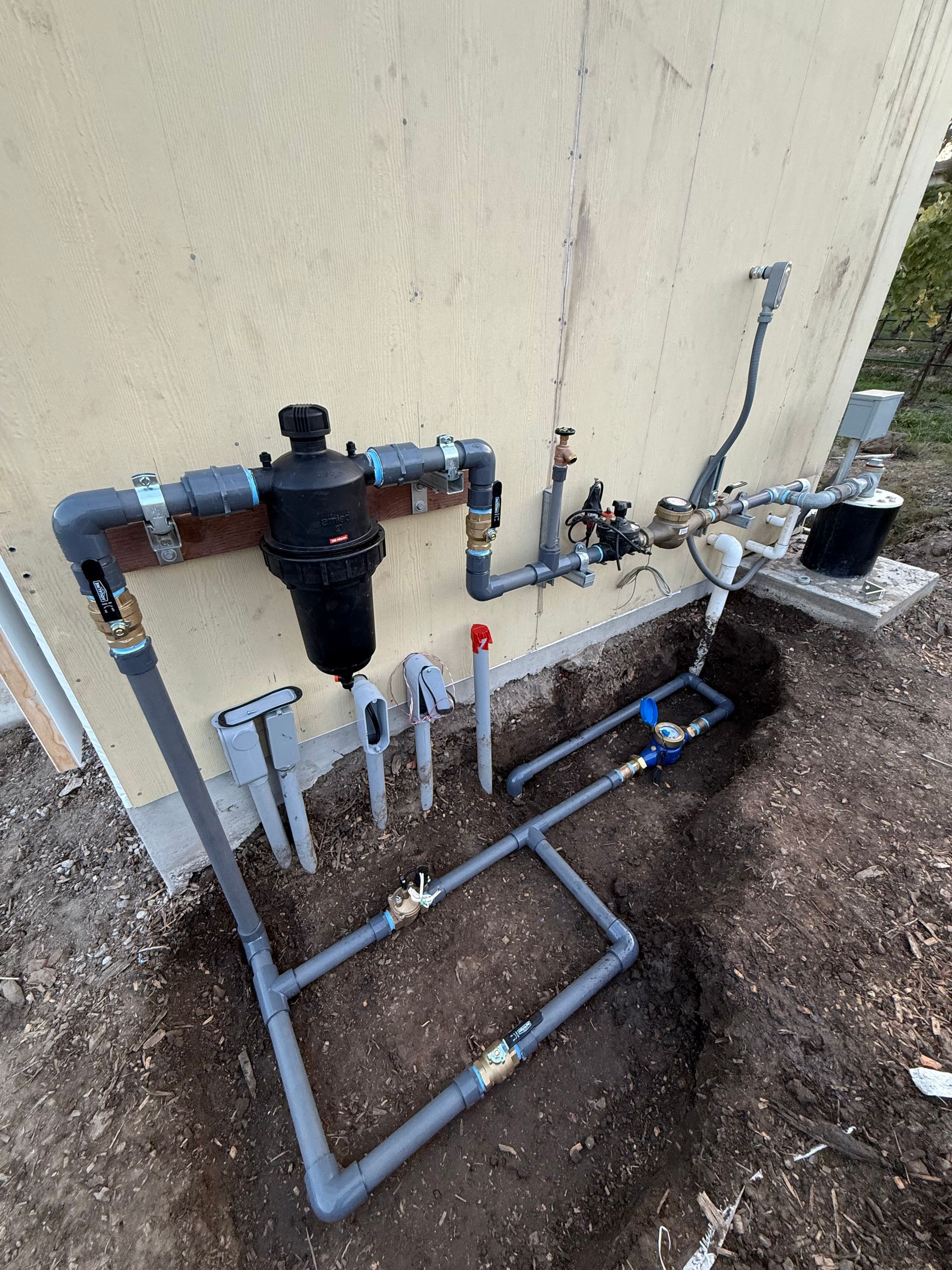 Outdoor water filtration and plumbing pipes mounted on a beige exterior wall.