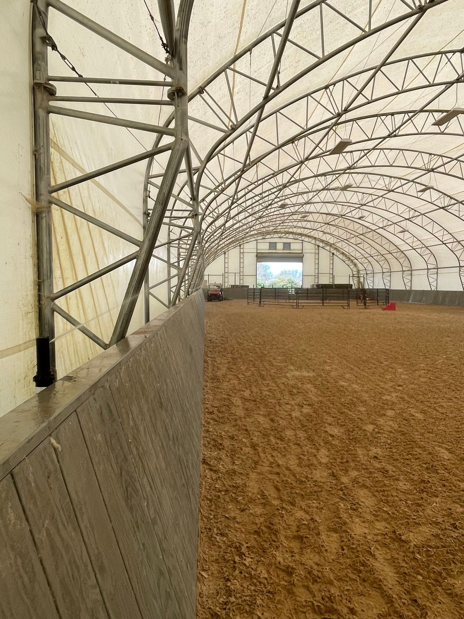 Indoor covered arena with dirt floor, metal trusses, and an open doorway at the far end