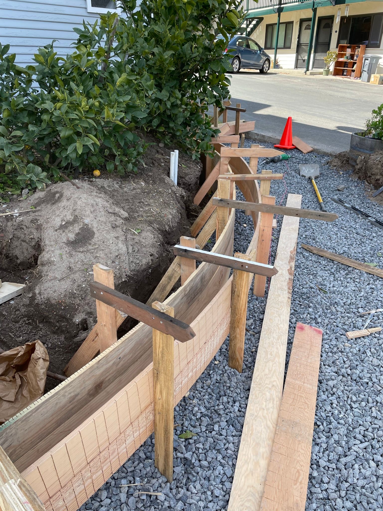 Wooden formwork along a gravel trench beside a bush, with an orange cone in the background.