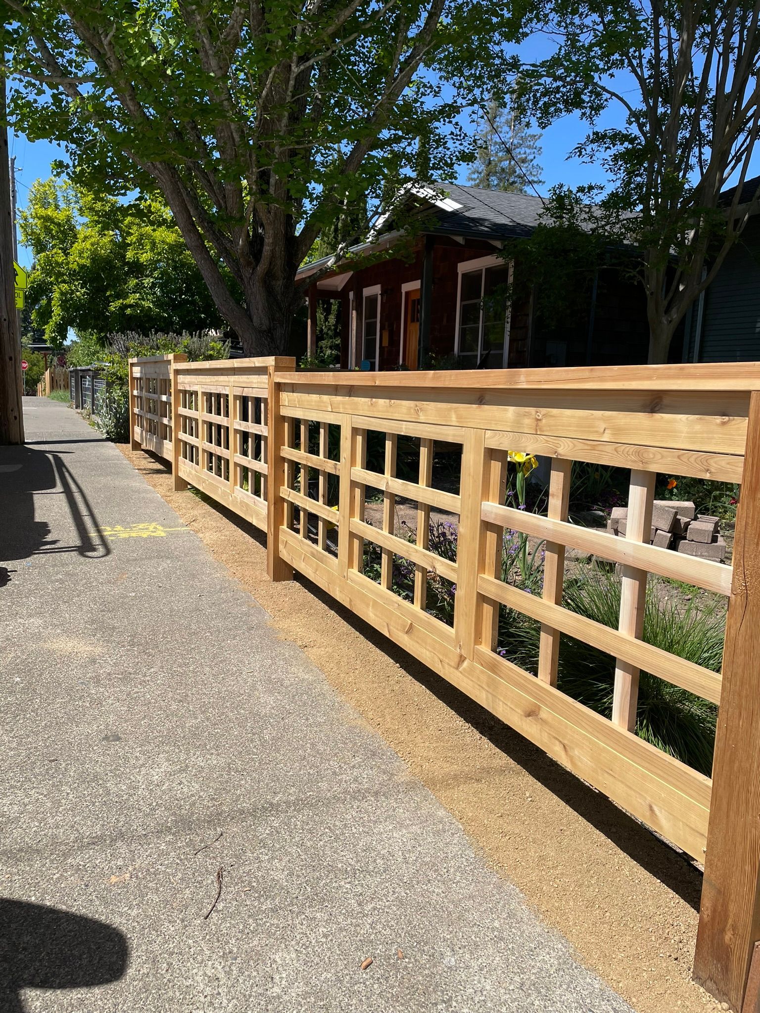 Wooden ramp railing along a gravel path beside trees and a small building in bright sunlight