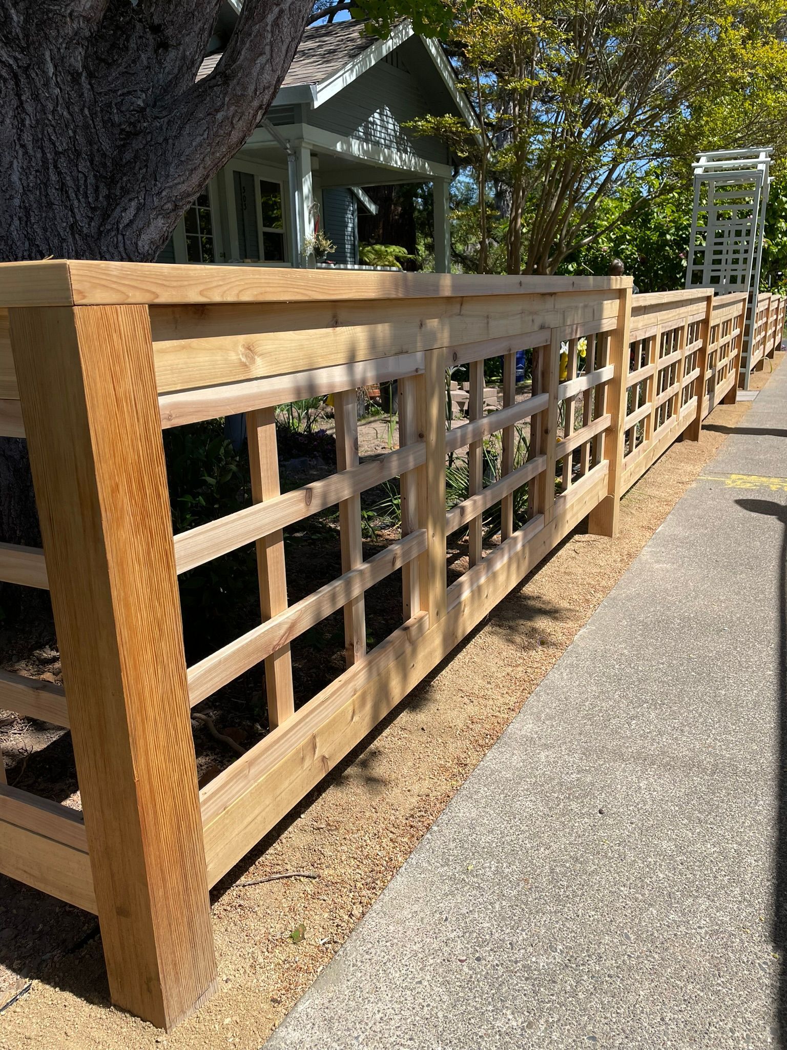 Wooden fence with lattice panels along a gravel path beside a large tree and house.