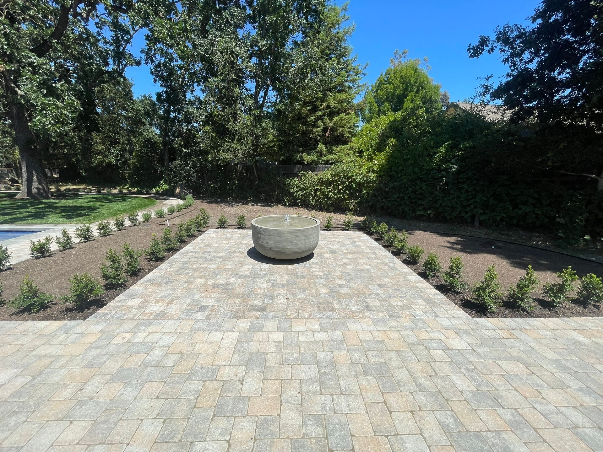 Stone planter centered on a paved courtyard, framed by low shrubs and tall green trees under a blue sky