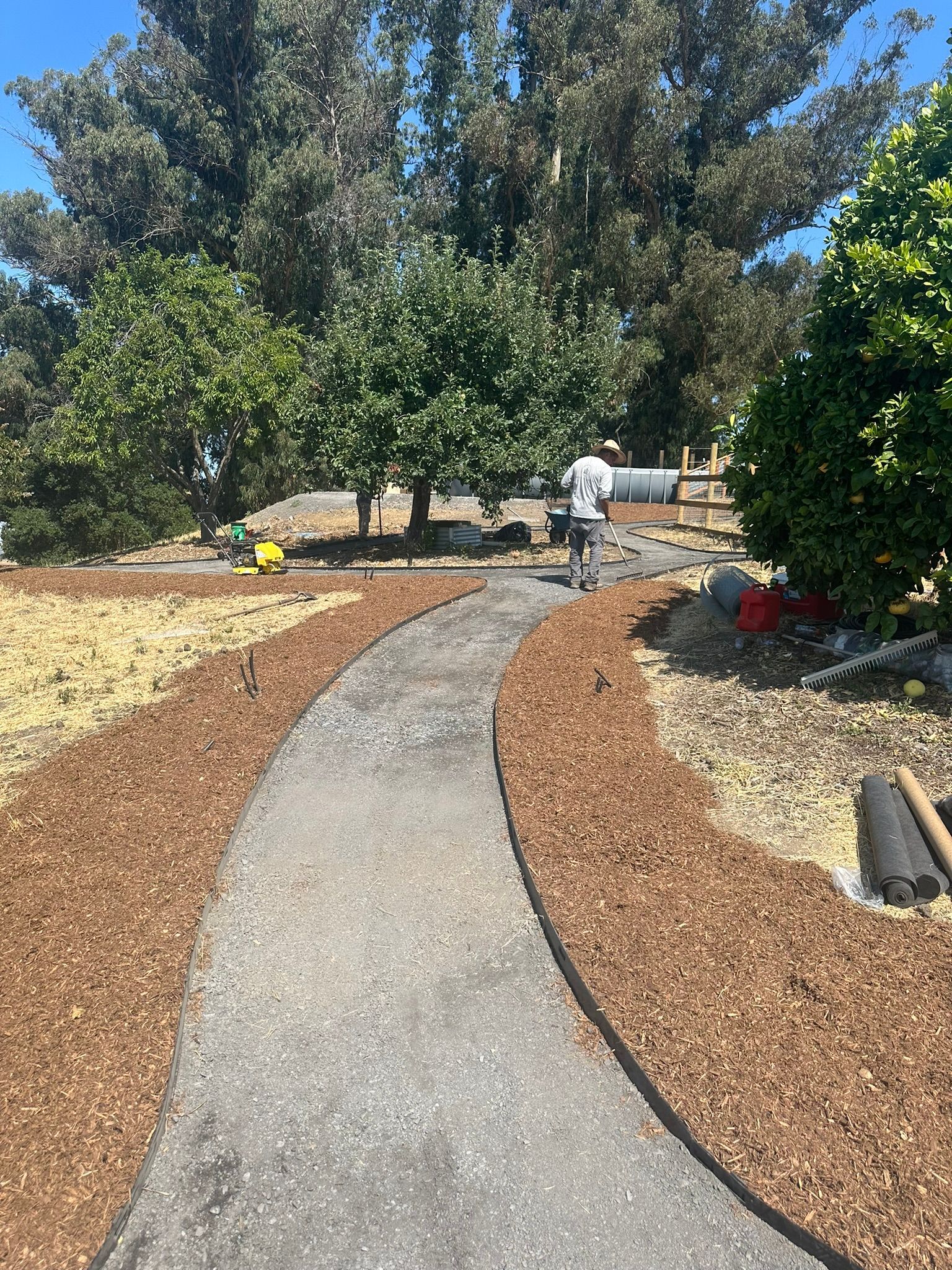 Curving gravel garden path through landscaped yard with trees and potted plants, worker in white in the distance