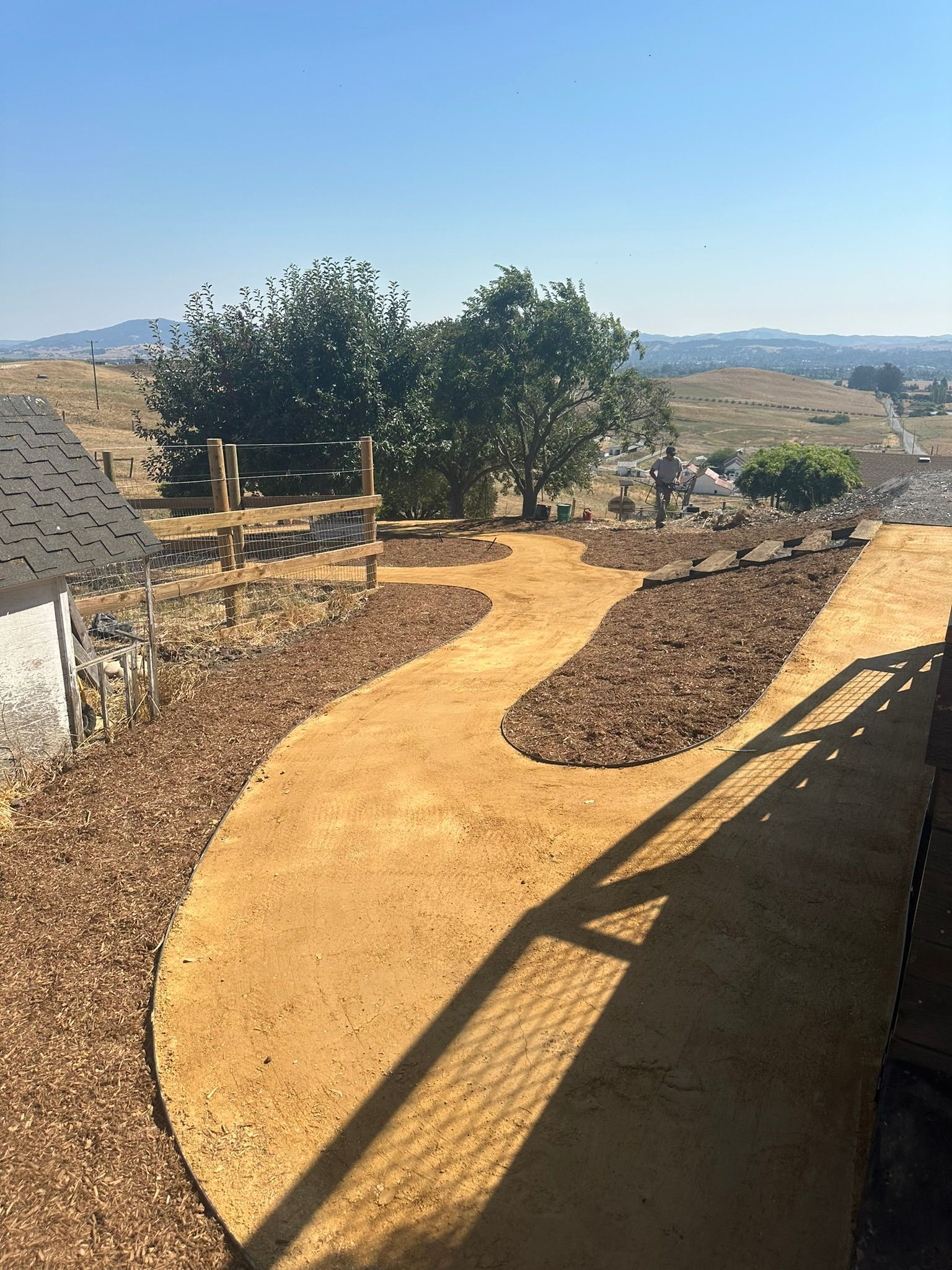 Curving tan gravel path through a dry garden with trees, benches, and hills under a clear blue sky