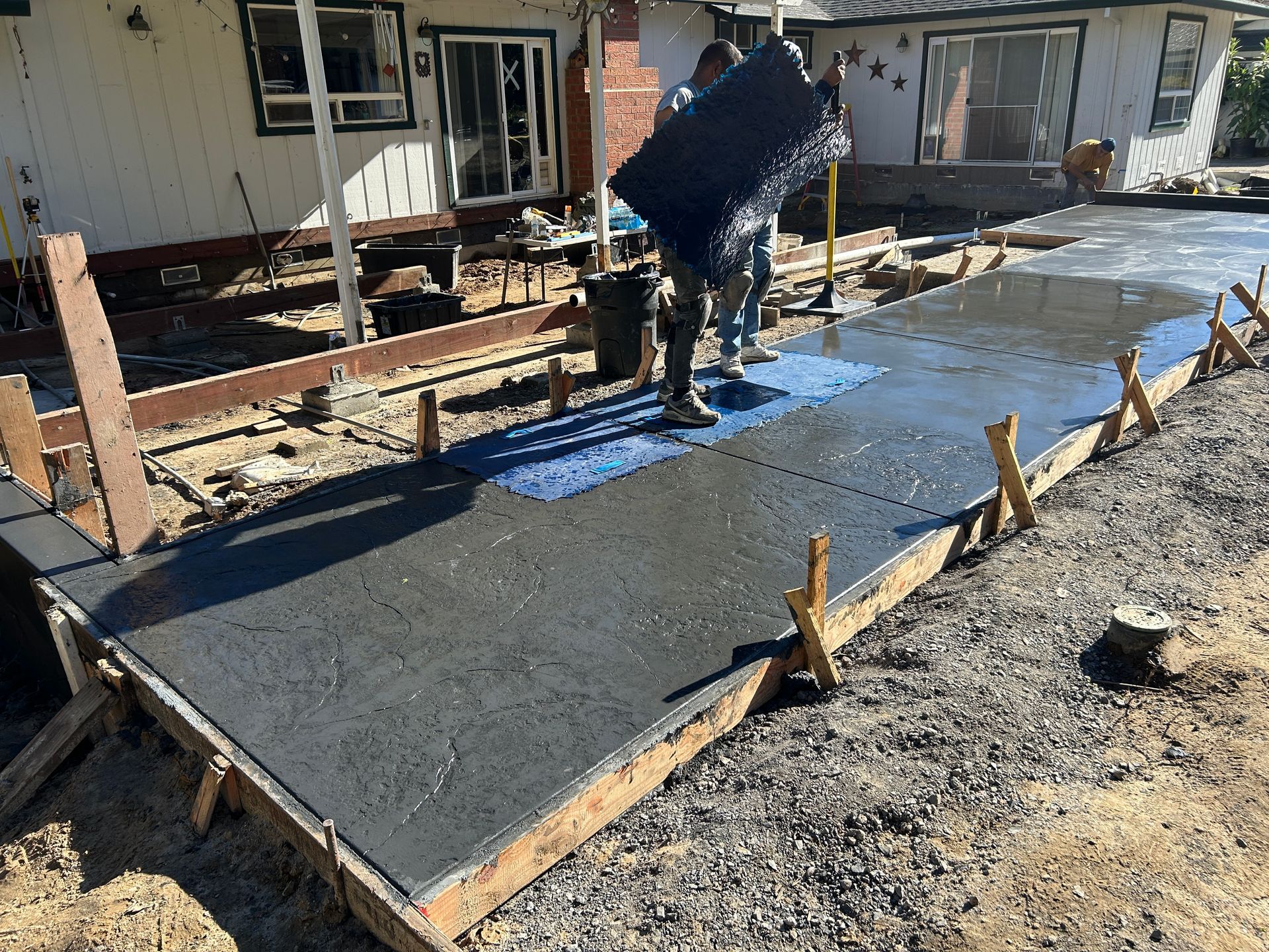 Worker pouring concrete on a new walkway beside a house, with wooden forms and wet cement.