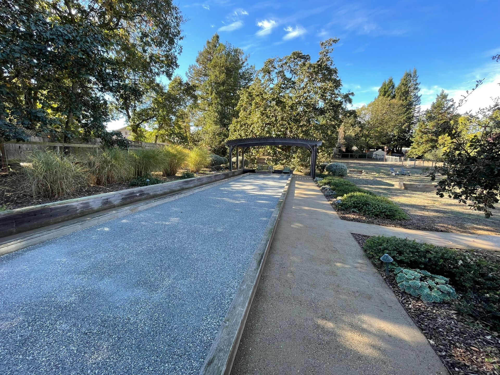 Gravel garden path curving through trees and shrubs toward a small bridge under a blue sky