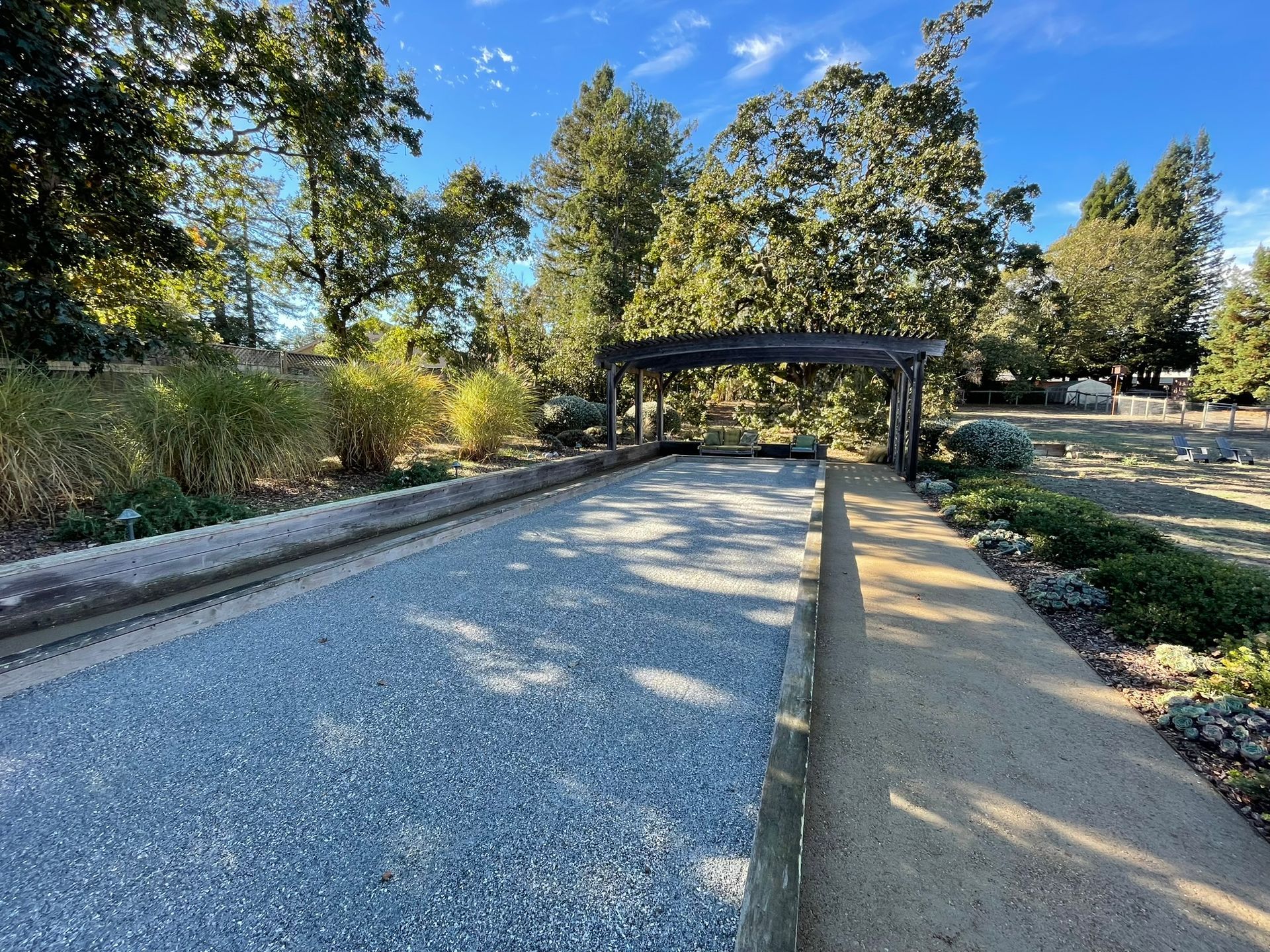 Gravel path with a central strip leading to a covered pergola in a sunny park, surrounded by trees and shrubs
