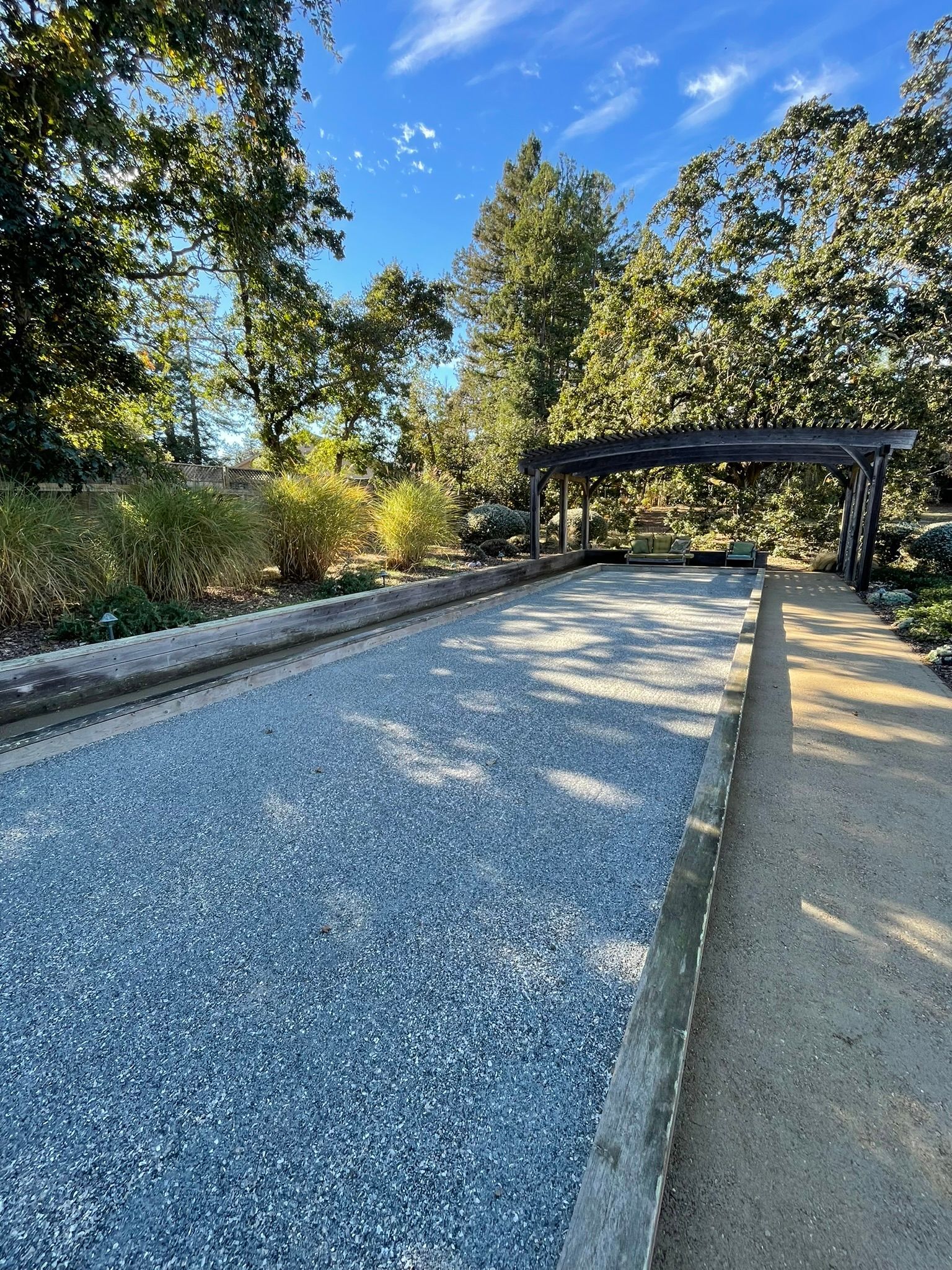 Gravel cornhole lane outdoors under a metal canopy, with trees and blue sky in the background