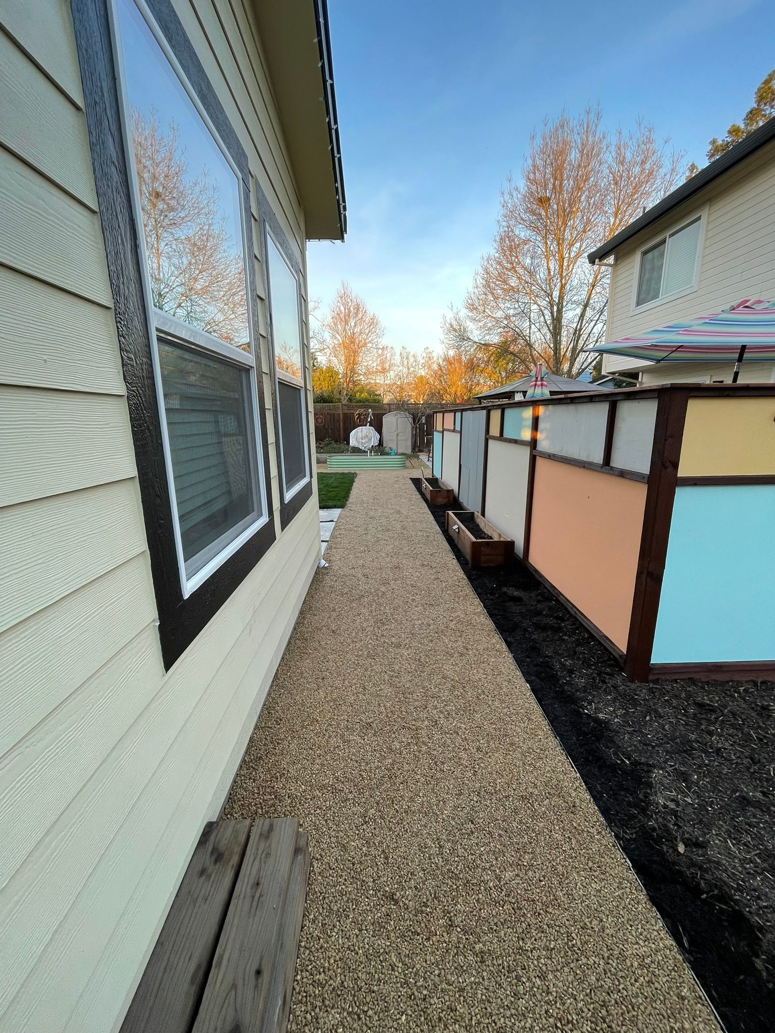 Gravel side yard between a white house and fence, with black edging and autumn trees in the background