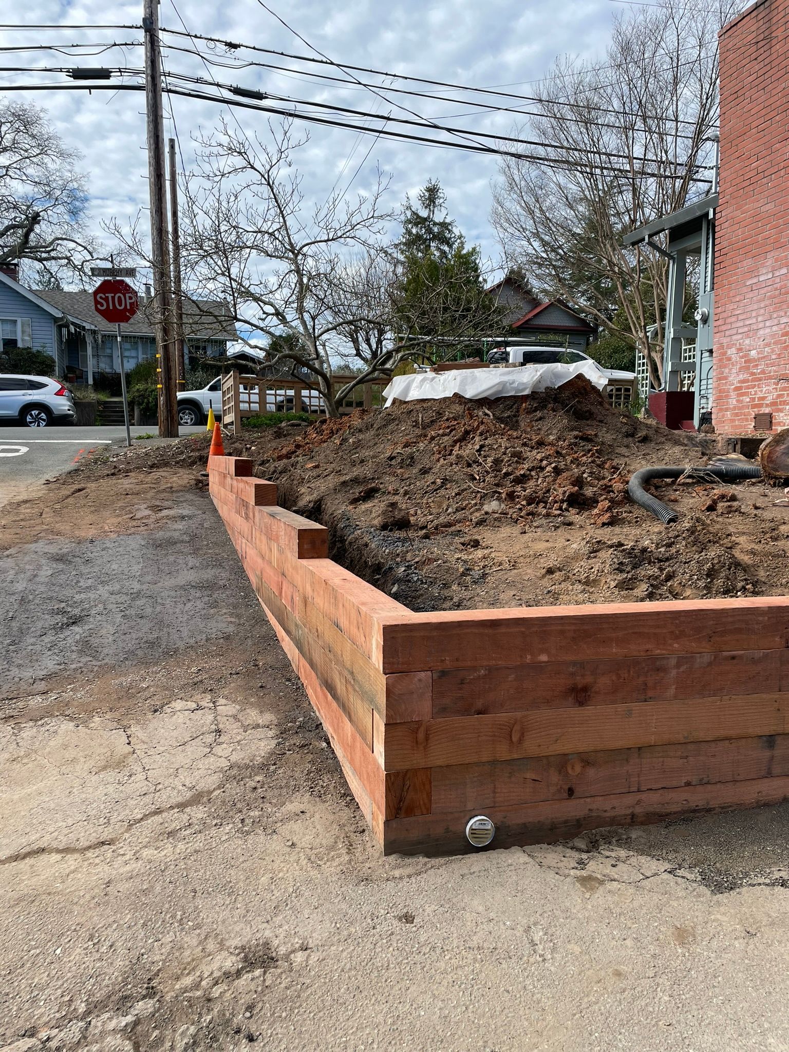 Wooden raised garden bed filled with mulch beside a sidewalk and street near a brick building