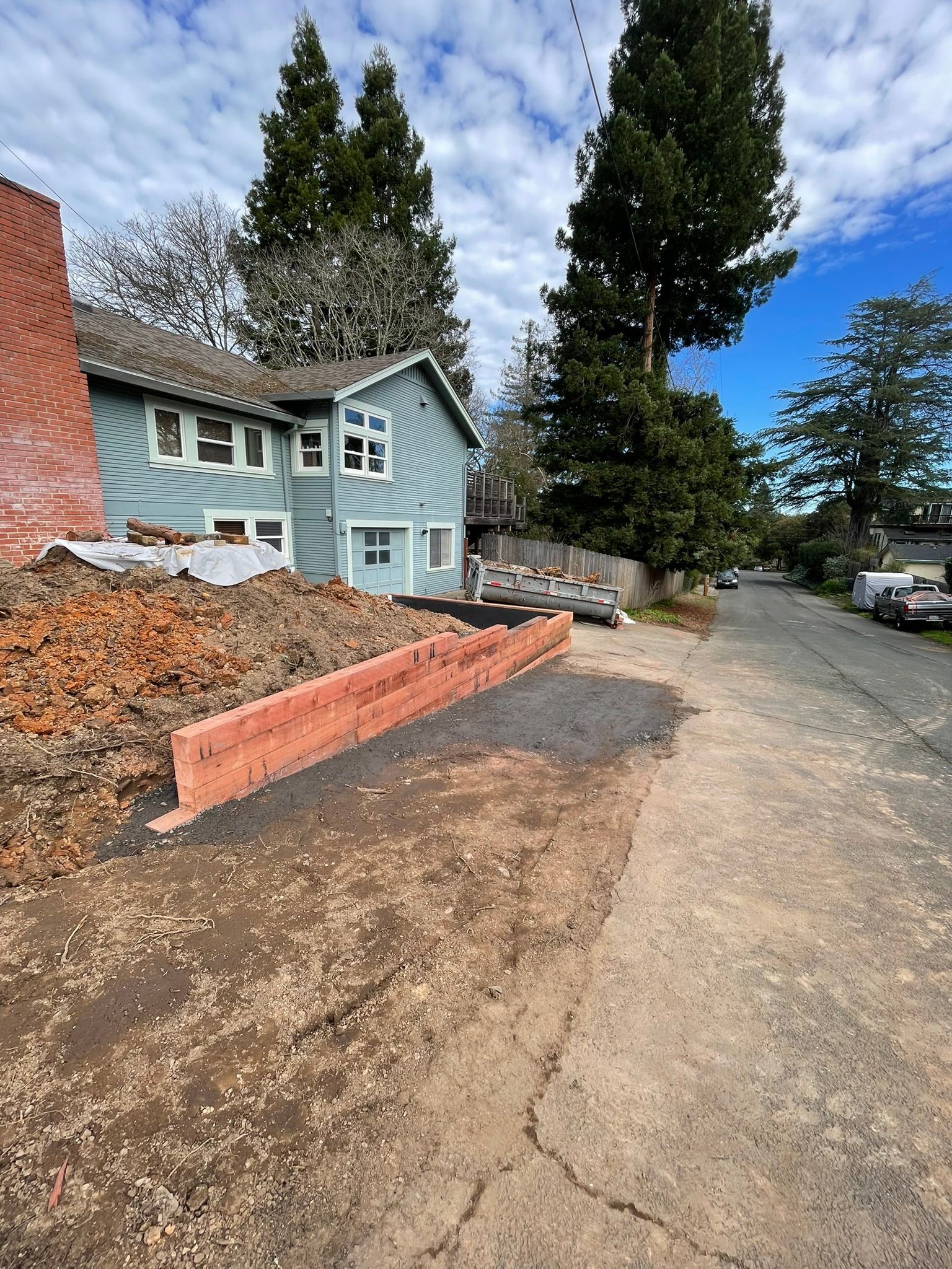 Residential driveway beside a light blue house, with tall evergreens, a brick retaining wall, and parked cars on a dirt road