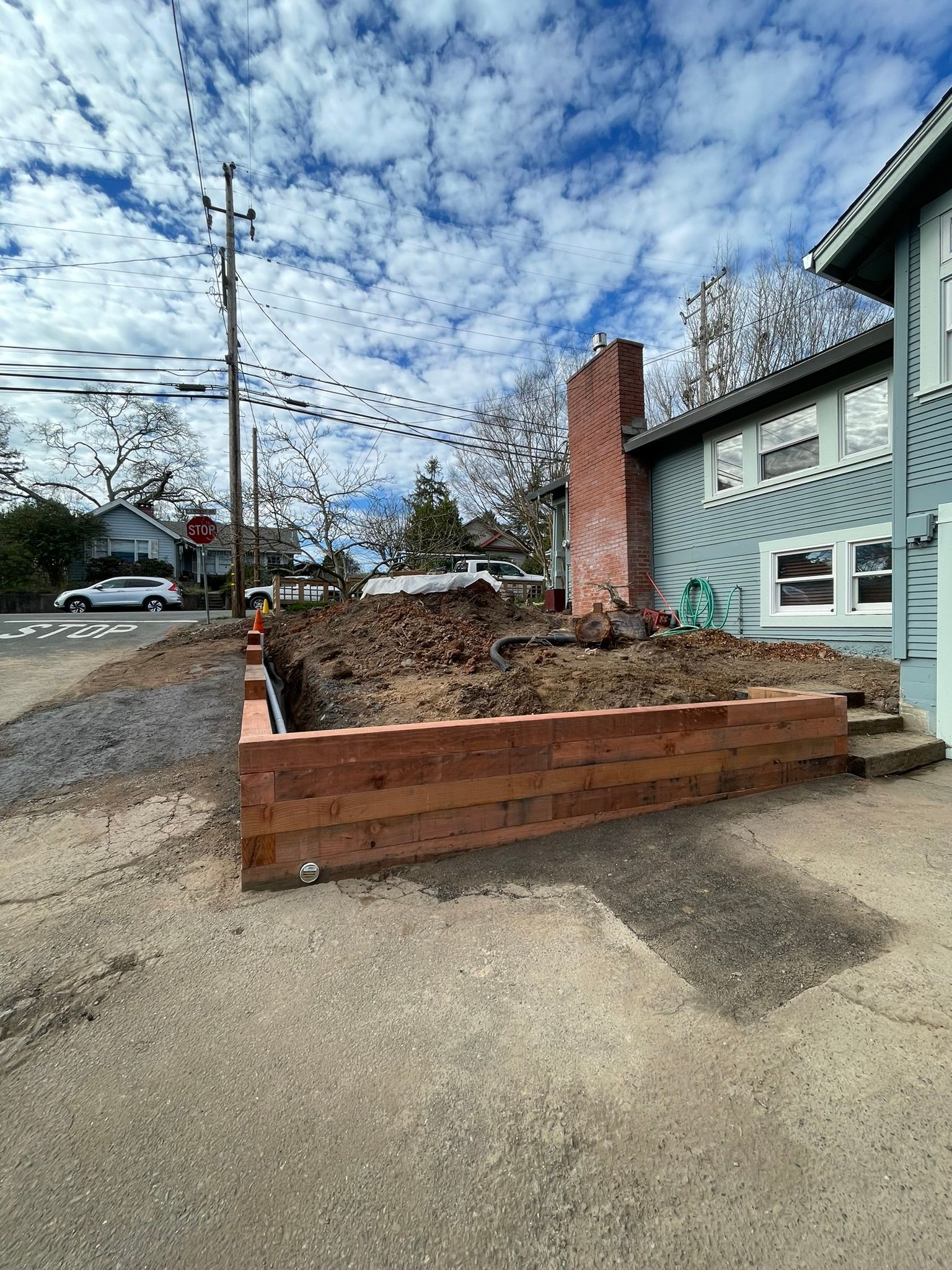 Street-side construction site with a brick retaining wall beside a light blue house under a cloudy sky