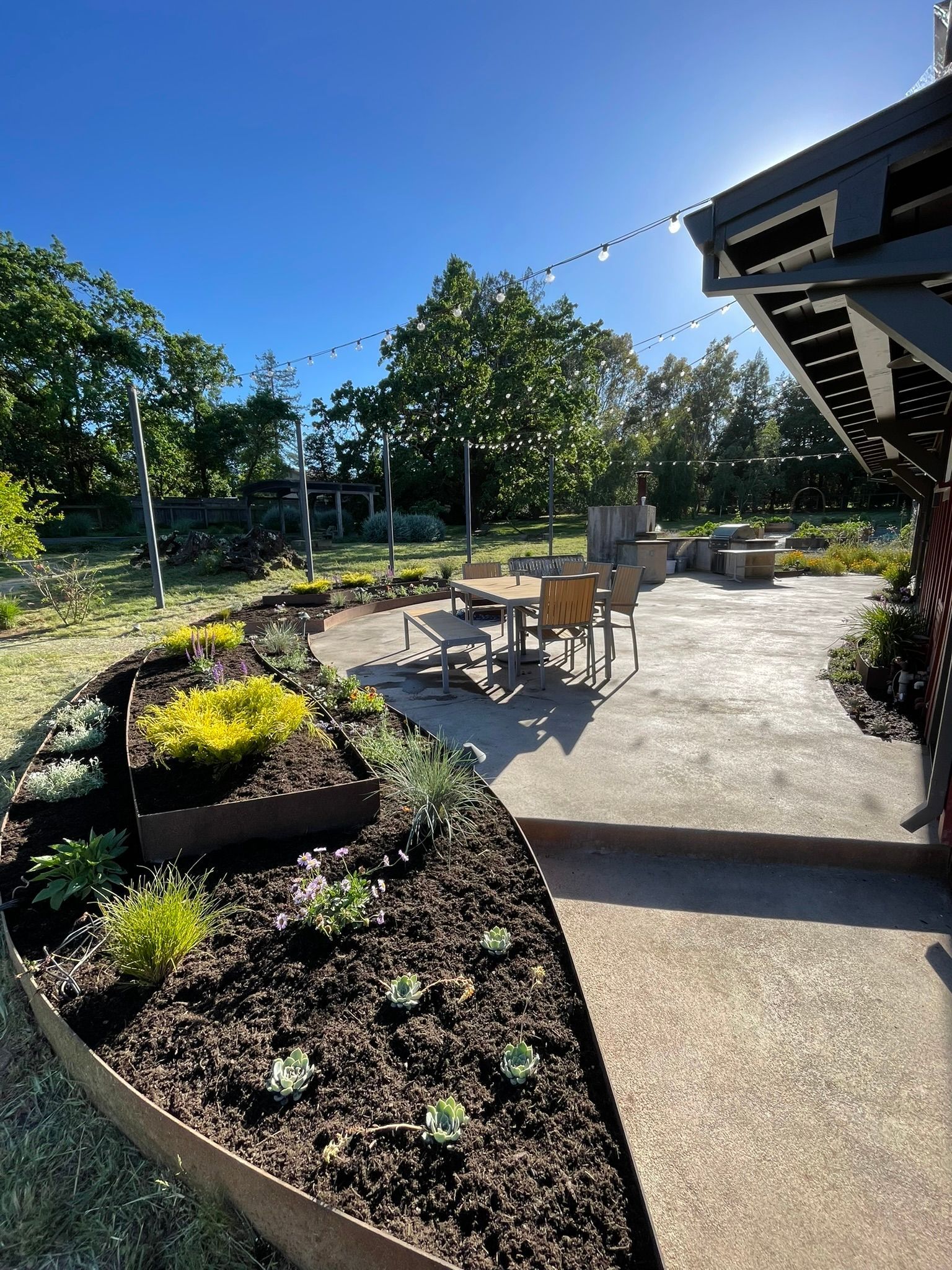 Sunny backyard patio with string lights, table and chairs, and raised flower beds beside a house