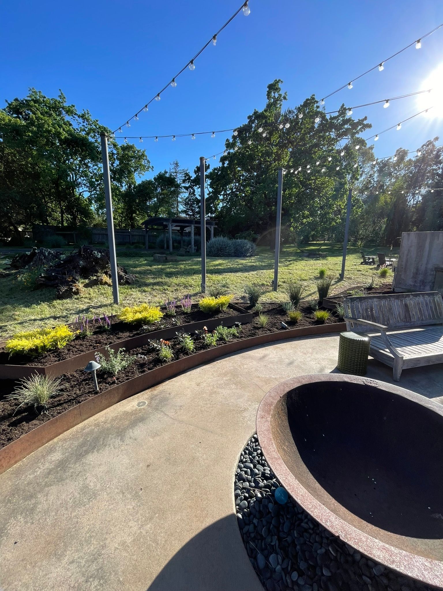 Sunny garden patio with string lights, flower beds, and a circular fire pit.