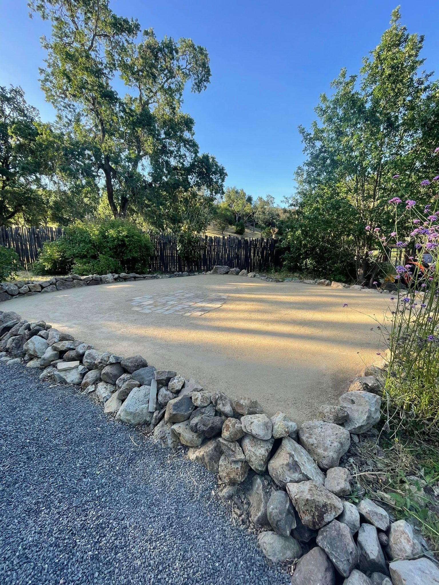 Gravel garden path bordered by rocks, leading to a circular sandy area with trees and blue sky behind it