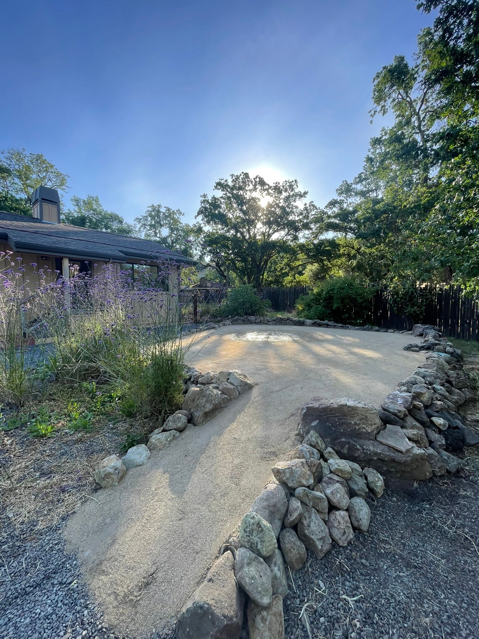 Curved gravel path beside a stone house, with rocks, trees, and bright sun in a garden setting
