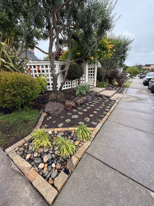Triangular rock garden beside a sidewalk with trees, shrubs, and a white fence on a wet street