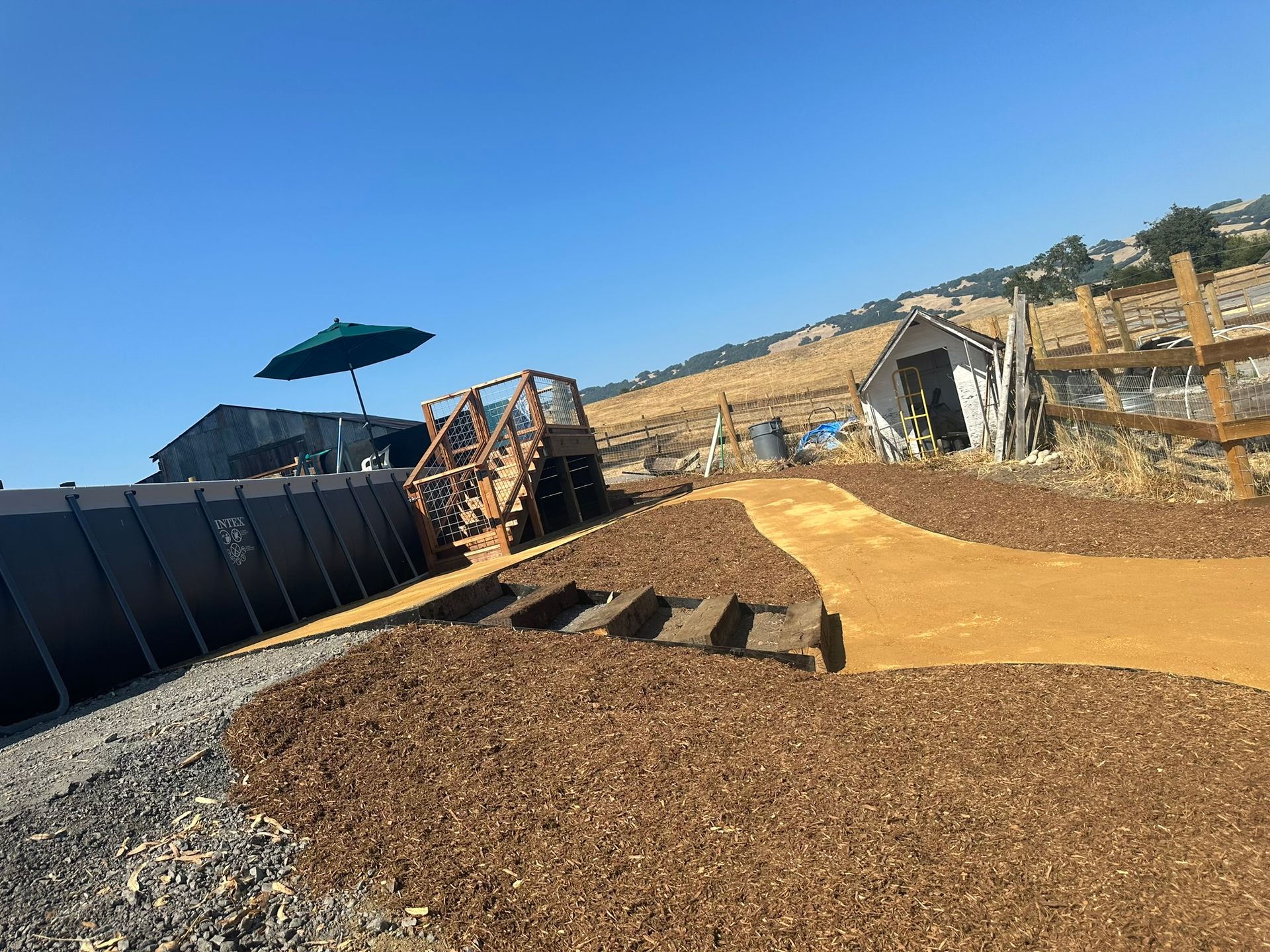 Outdoor dirt play area with a small wooden ramp, stairs, and a shaded umbrella under a clear blue sky
