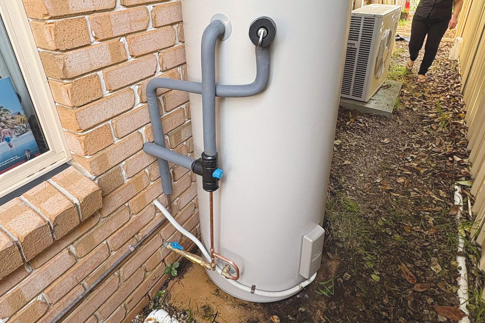 An outdoor cylindrical hot water heater with insulated plumbing pipes against a brick wall.