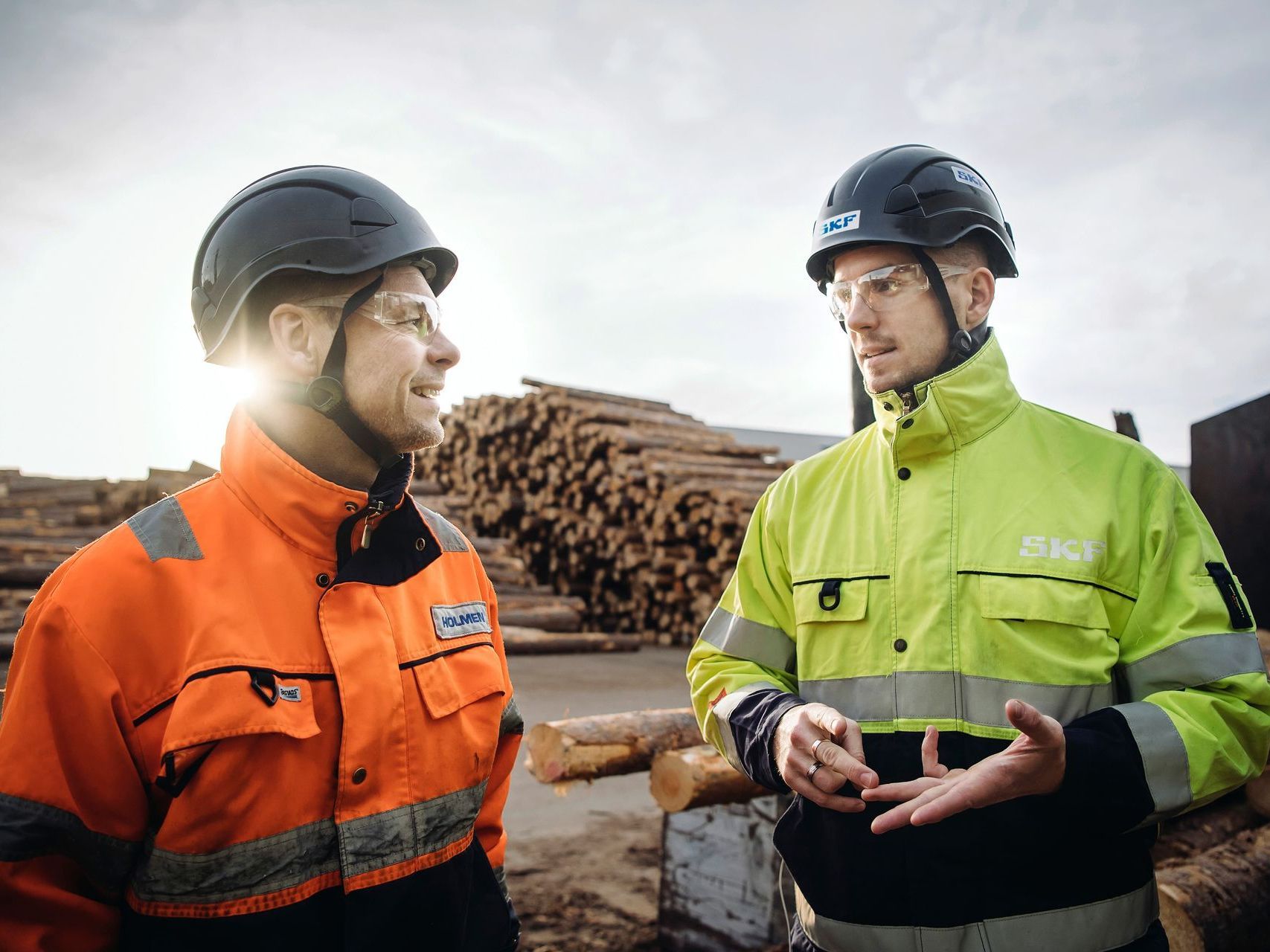 Två arbetare i hjälmar och varseljackor pratar på en såggård, med staplade stockar bakom sig.