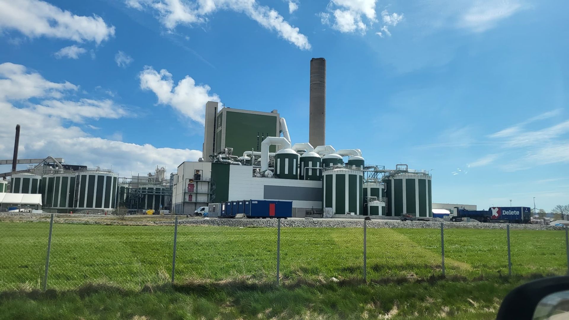 Industriellt kraftverk med hög skorsten under en blå himmel, sett bortom ett gräsbevuxet staket.