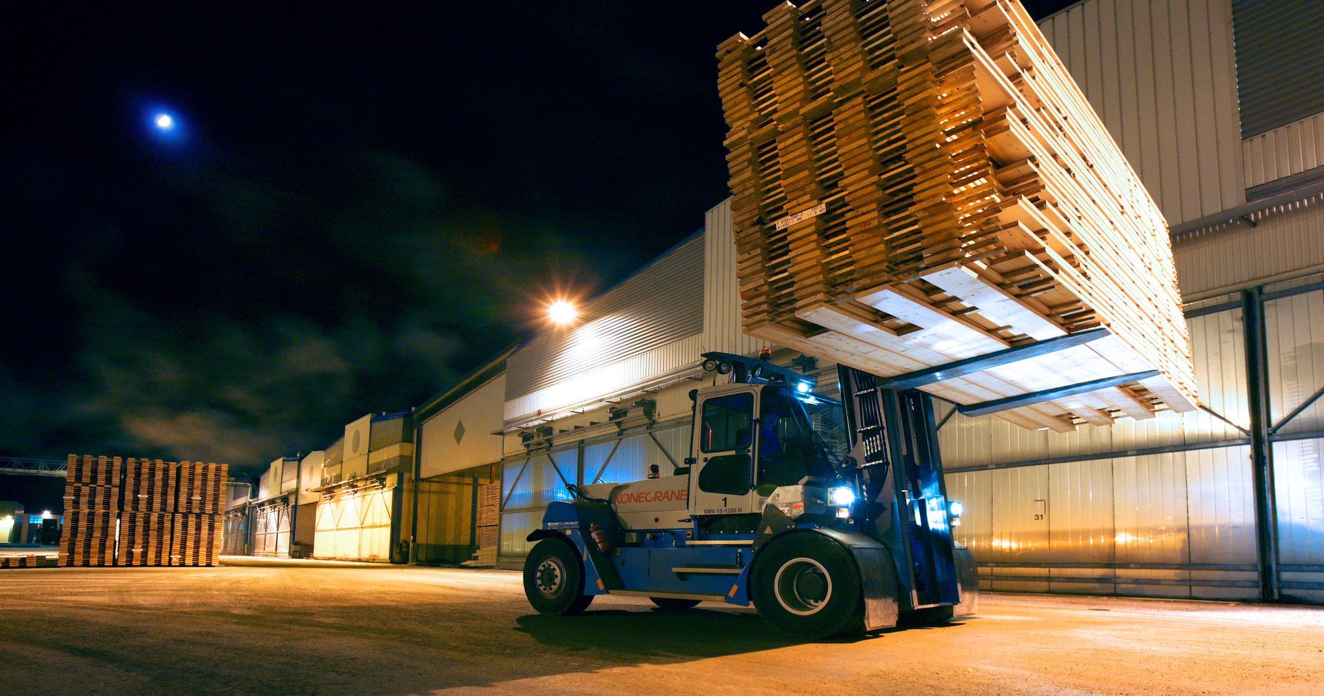 Gaffeltruck som staplar stora träpallar på natten utanför ett industrilager.