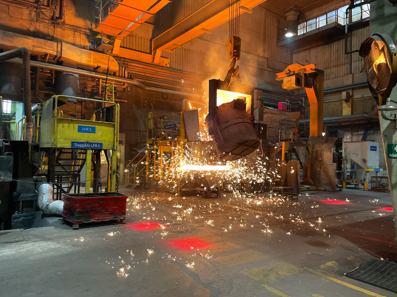 Smält metall hälls i ett industriellt gjuteri, med ljusa gnistor och orange upplyst maskineri.