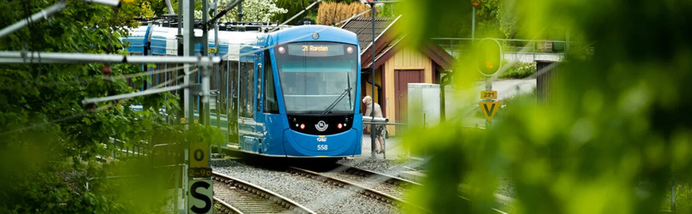 Blå spårvagn på böjda spår i en grön stadsmiljö, närmar sig en station