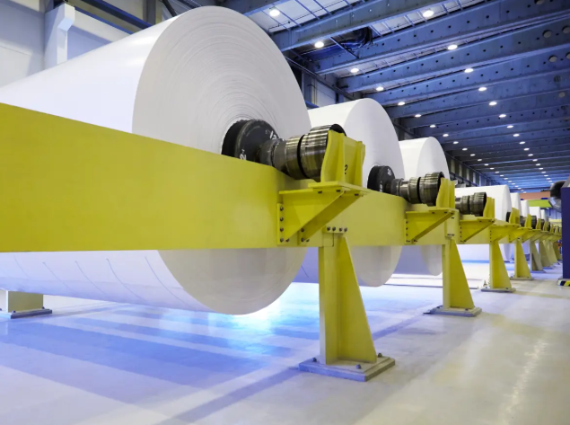 Stor gul industrimaskin med överdimensionerade vita rullar i en fabrik eller pappersanläggning