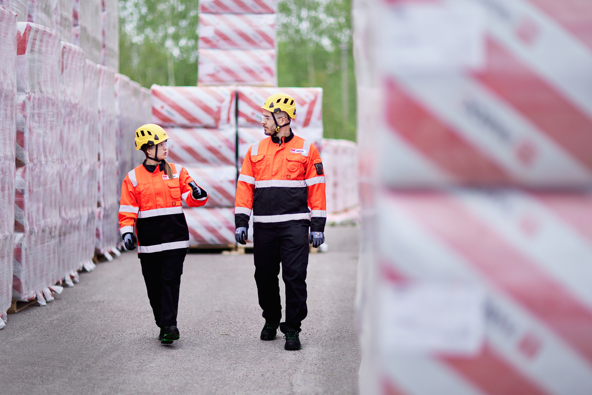 Två arbetare i orange säkerhetsvästar och hjälmar går mellan staplad last på en industrigård