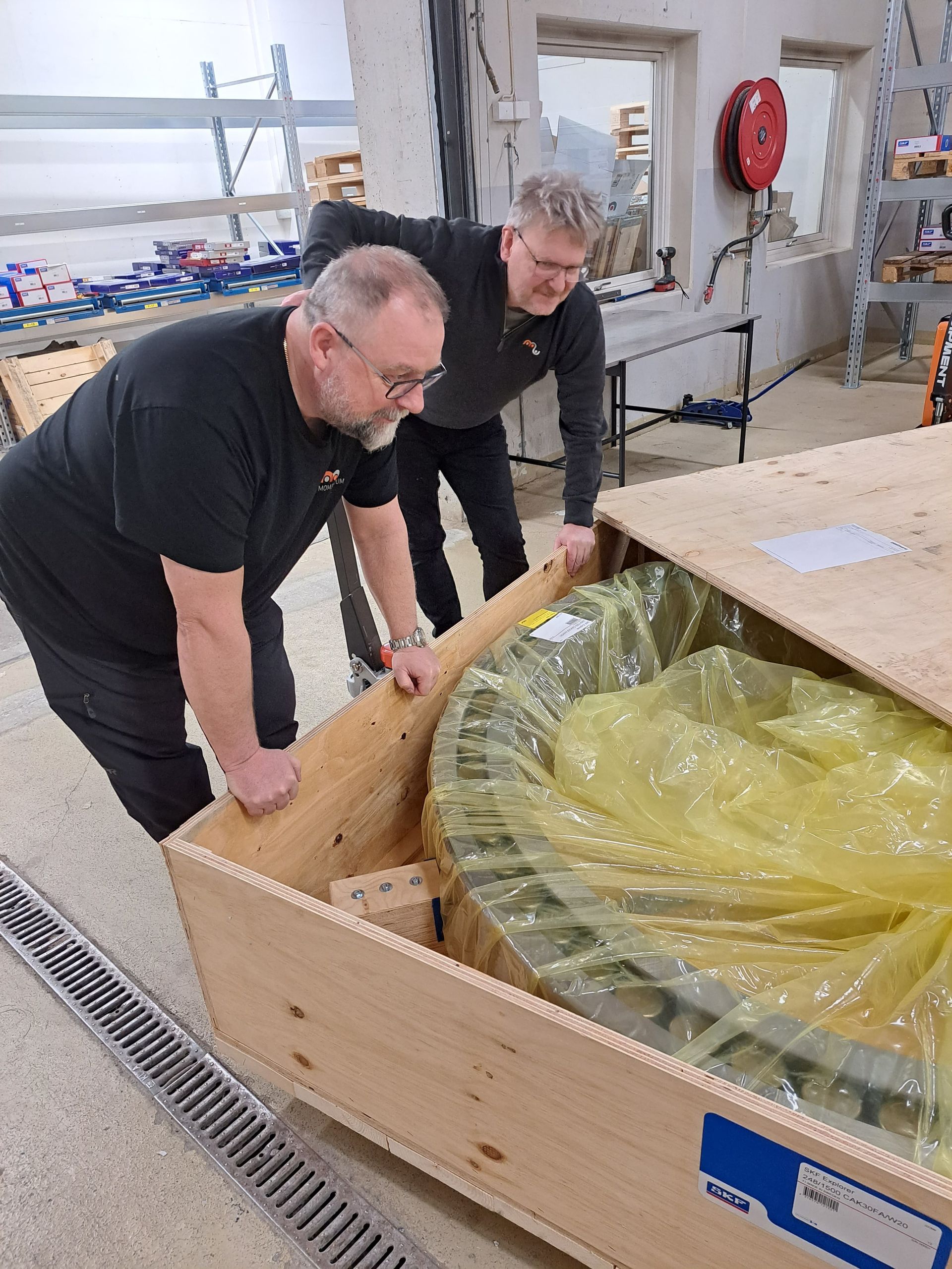 Två arbetare lyfter en stor trälåda insvept i gul plast i en verkstad.