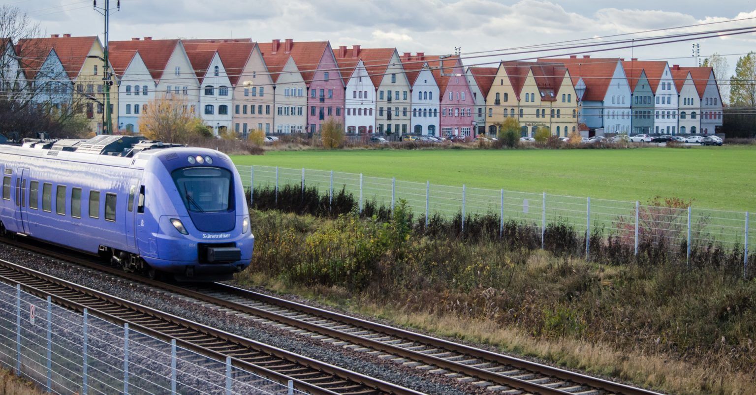 Blått passagerartåg på spår bredvid ett grönt fält och en rad av färgglada hus